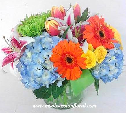 Bright mixed bouquet with blue hydrangeas, orange gerberas, yellow roses and lilies in a glass vase