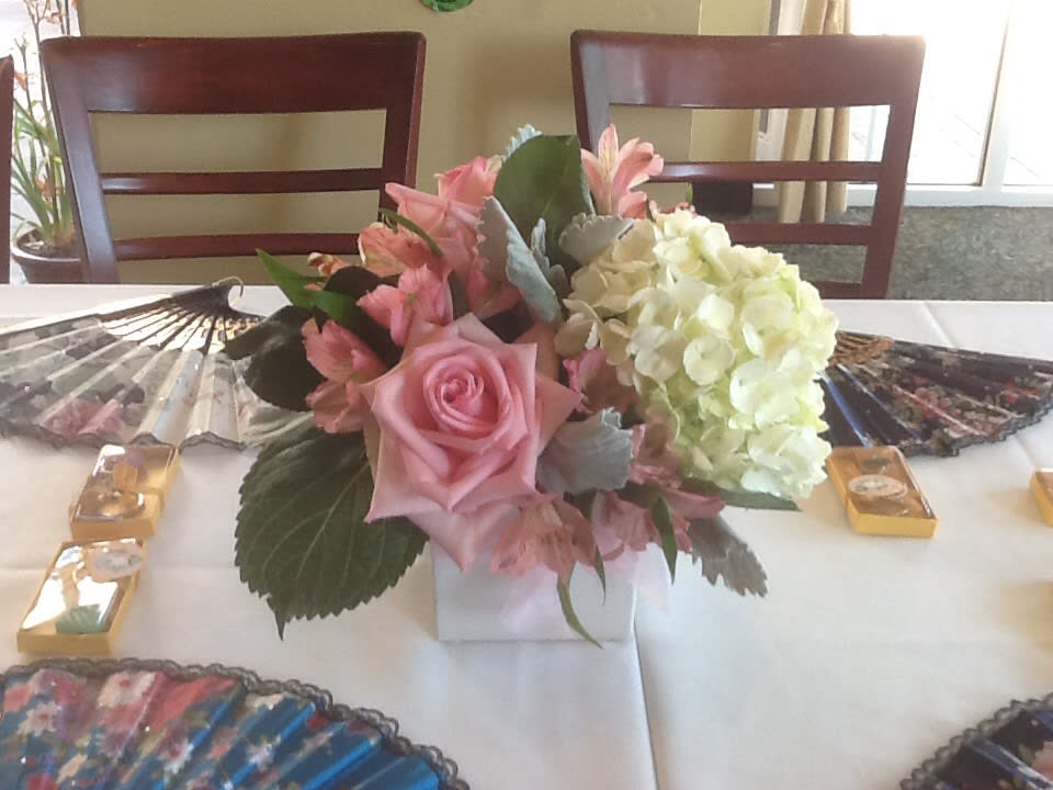Event centerpiece with pink roses and white hydrangeas in a white cube vase on a table with fans.