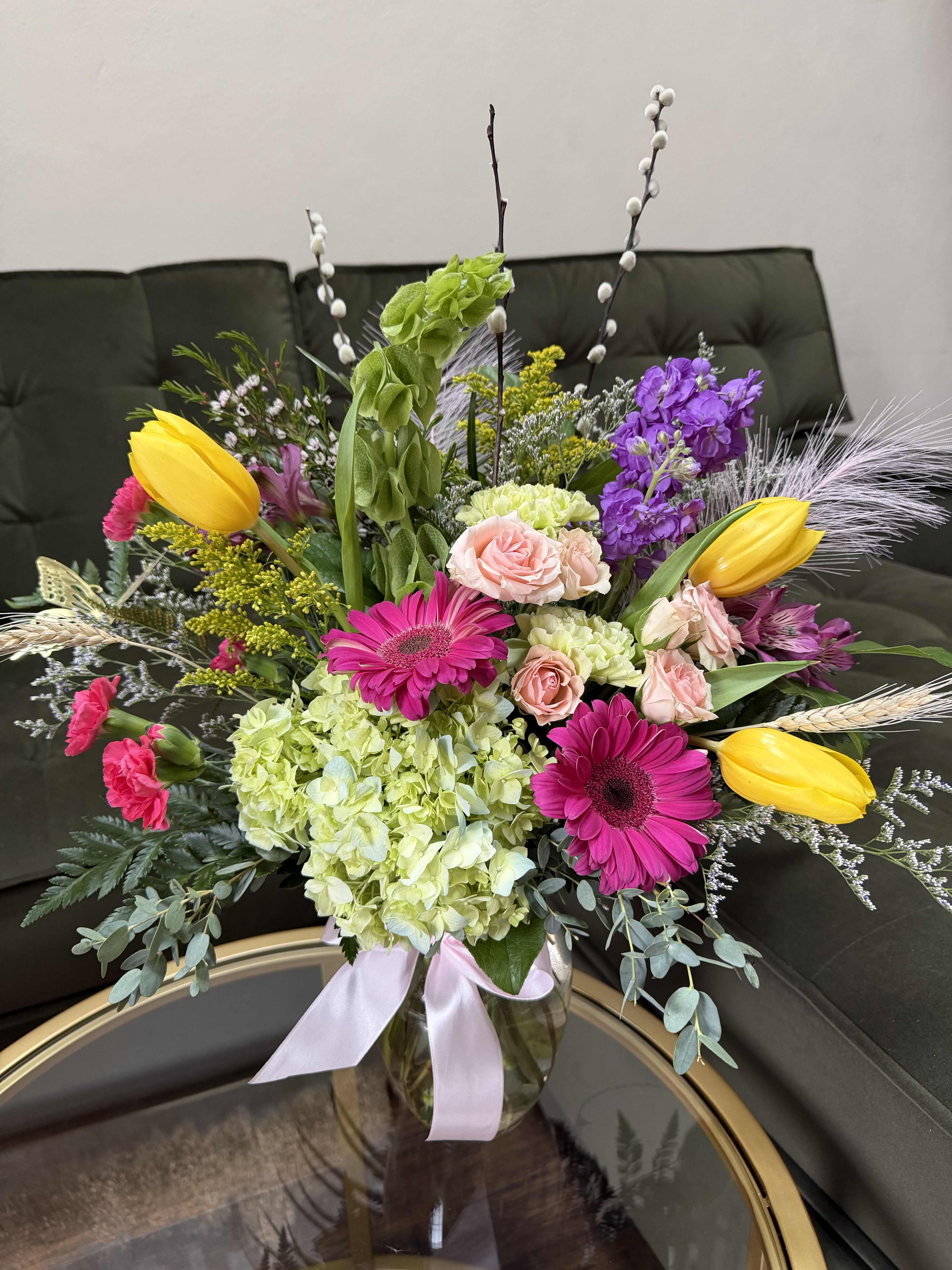 Radiant Spring Reverie - A vibrant, garden-inspired composition that feels both refined and effortlessly abundant. This arrangement features luminous yellow tulips, velvety stock, gerbera daisies, and delicate blush spray roses nestled among lush hydrangea. Accents of fresh spring foliage, textural pussy willow branches, and airy wisps of ornamental grass create movement and dimension, while subtle touches of lavender and soft greenery add depth and elegance.   Designed in a clear glass vase finished with a soft satin ribbon, this piece captures the essence of modern luxury—fresh, joyful, and artfully untamed. Perfect for celebrating new beginnings, elevating interiors, or gifting a statement of sophisticated beauty, as well as Easter and Spring seasonal gifts.