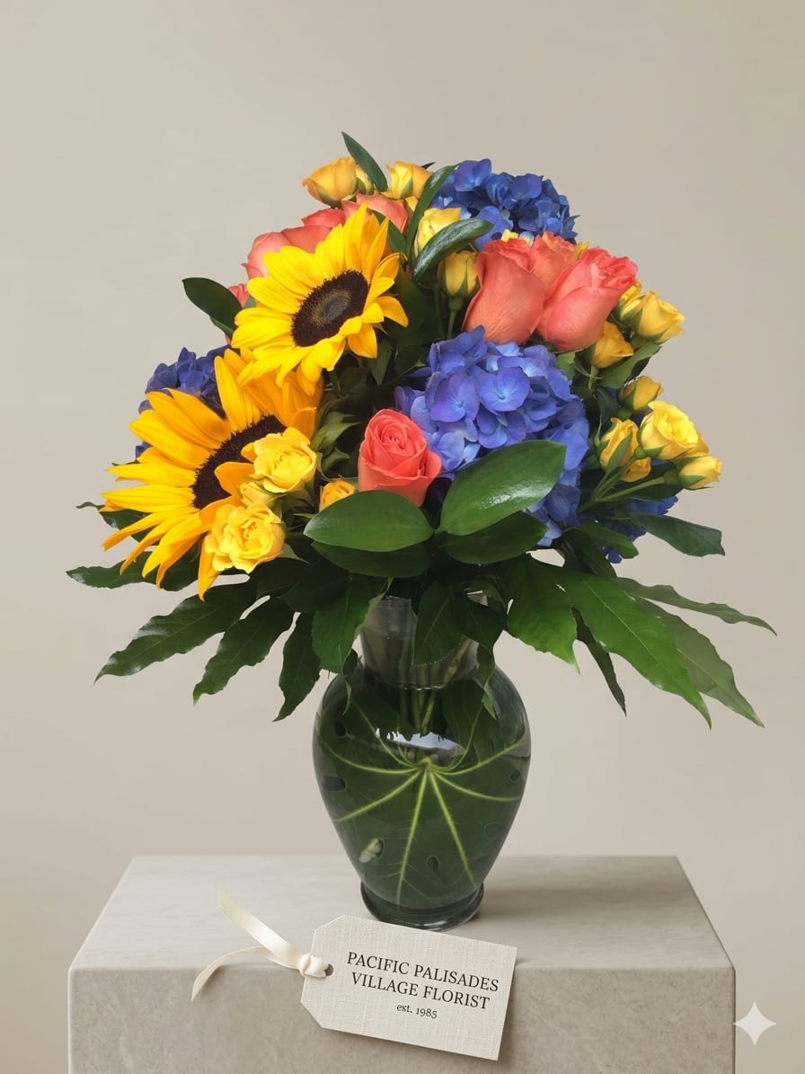 Fields of Gold - A bright, cheerful arrangement that blends sunny yellow sunflowers with soft peach roses and clusters of blue hydrangea, accented by delicate yellow spray roses and lush greenery. The overall look is fresh and uplifting, with a balanced mix of bold focal blooms and soft, rounded textures arranged in a leafy glass vase.
