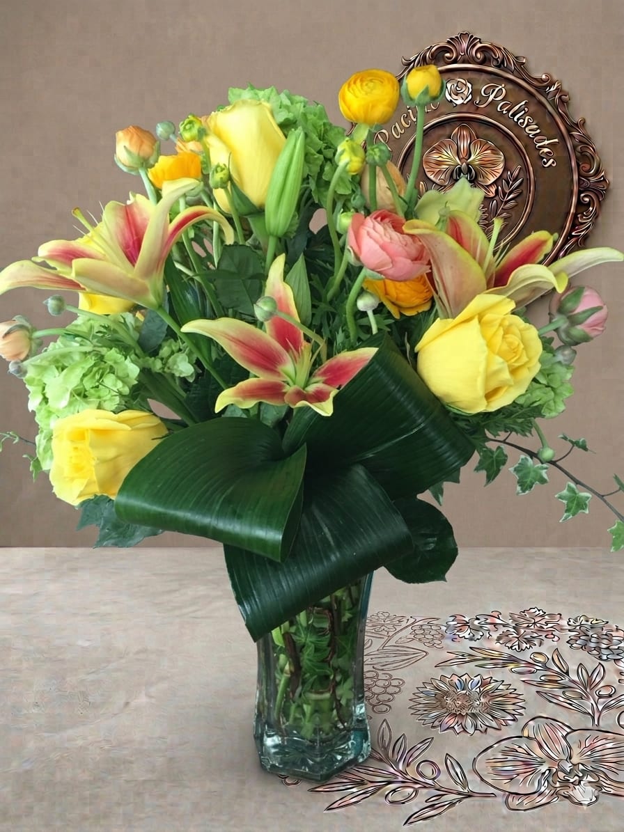 Hugs and Kisses - A lively, garden-inspired arrangement featuring sunny yellow roses and striking bi-color lilies, complemented by soft peach ranunculus and bright yellow accents, all layered with lush greenery and bold sculptural leaves. The design feels vibrant and full of movement, blending fresh spring tones with a modern, structured presentation.