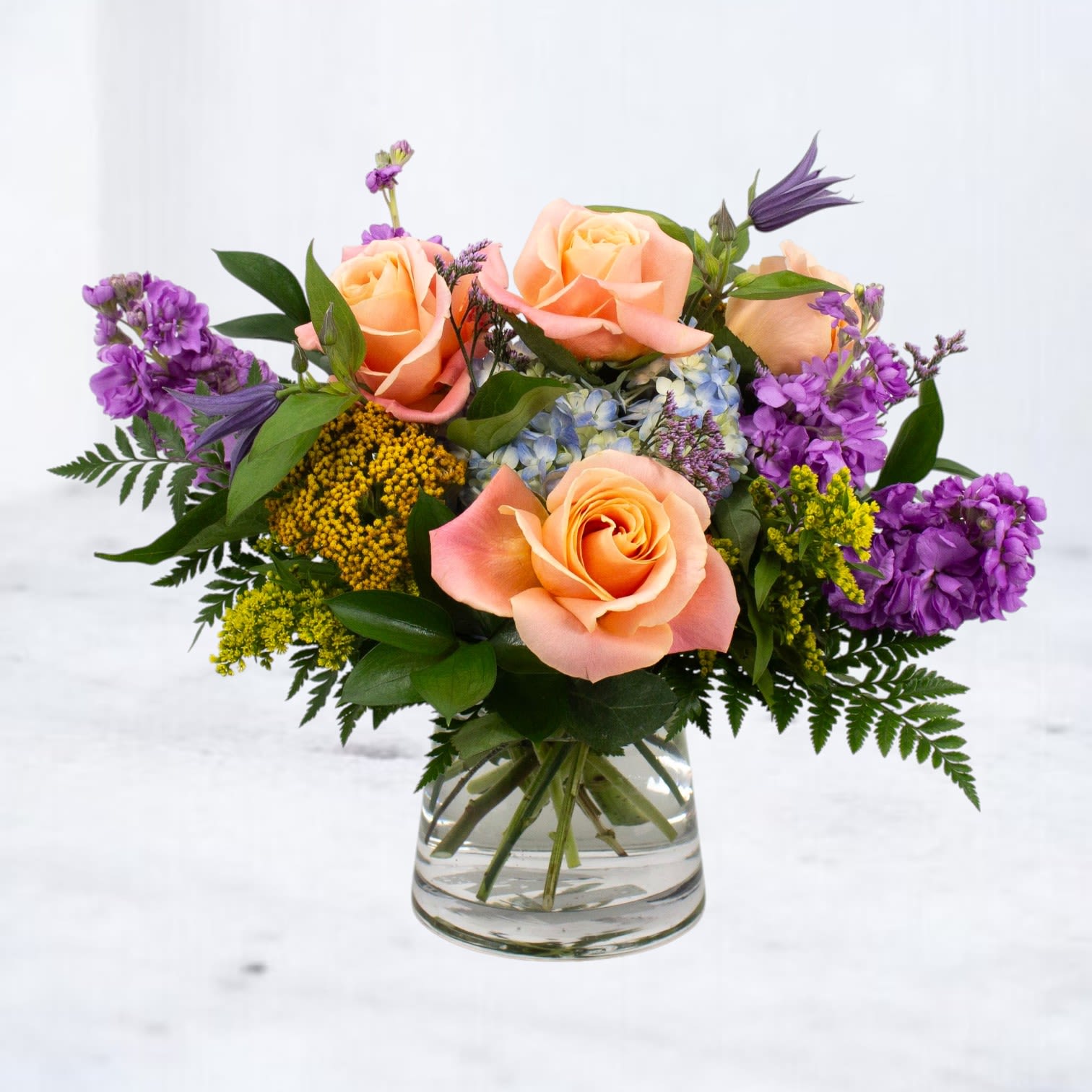 Rhapsody - A bold and unexpected harmony of color, Rhapsody is a striking floral arrangement that brings together the peachy warmth of roses with the moody richness of purple stock, blue hydrangea, golden yarrow, and wisps of lavender. Designed in a sleek clear glass vase, this arrangement dances with contrast and movement—like a rhapsody at dusk.  Perfect for birthdays, celebrations, or just because, this bouquet is a show-stopping gift for anyone who loves a little drama and beauty.