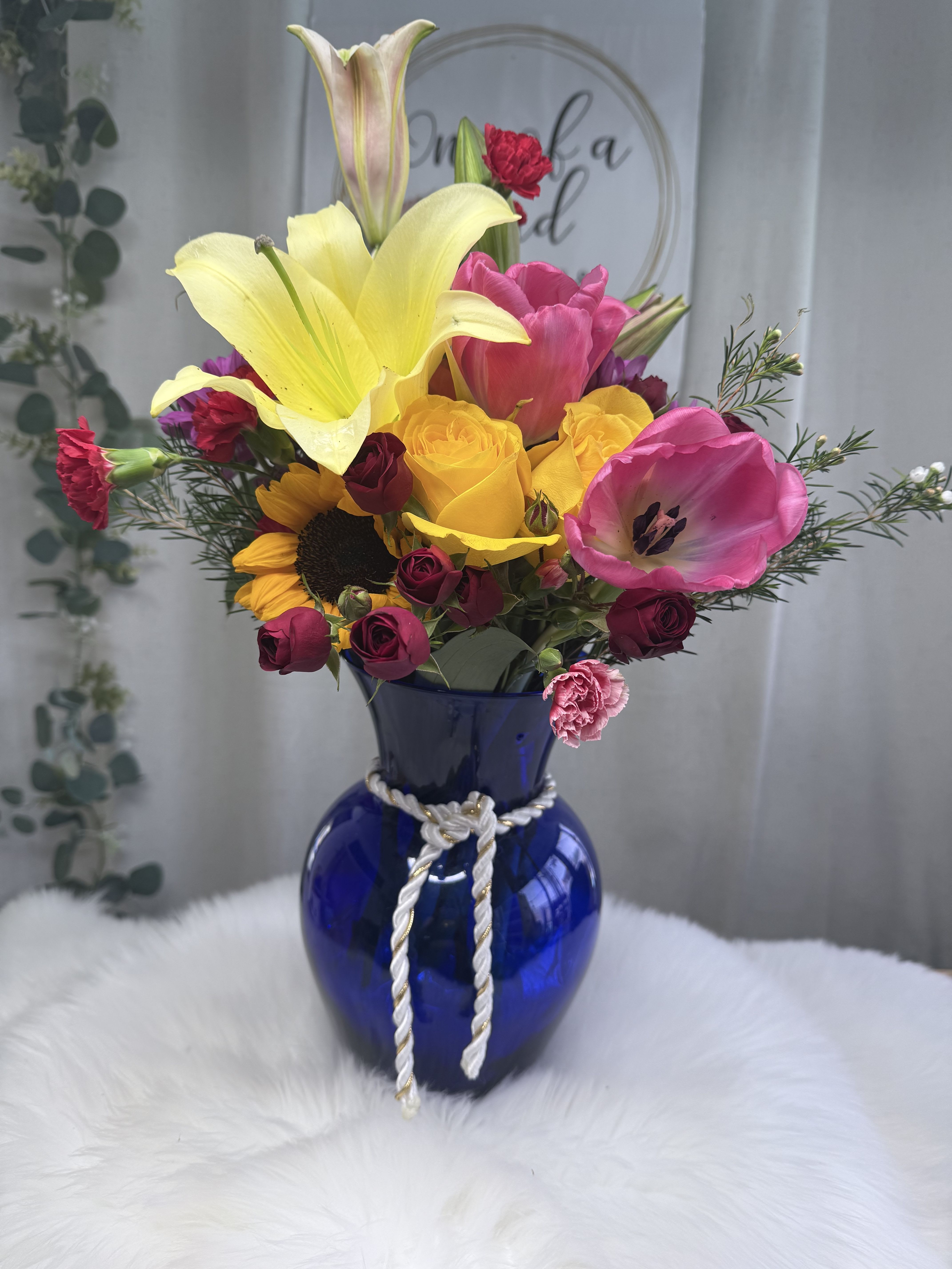 Golden Bloom Harmony - A lively and elegant floral arrangement bursting with seasonal color and charm. This design features a stunning combination of fresh blooms.  Arranged in a deep blue glass vase with a decorative rope accent, this bouquet brings a perfect balance of brightness and sophistication—ideal for celebrations, thank-you gifts, or simply adding beauty to any room.  Flower varieties may vary depending on seasonal availability while maintaining the same color palette and overall style.