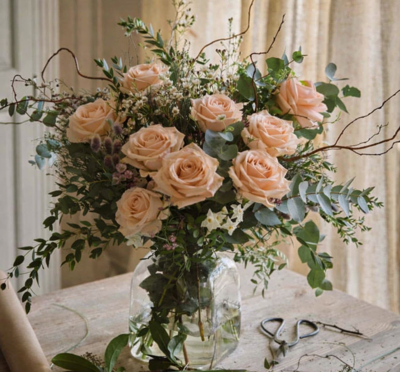 Peachful Chamber - A soft, romantic composition that feels like a quiet moment in bloom. Peachful Chamber features lush peach roses nestled among airy greenery and delicate touches of wax flower, creating a naturally graceful silhouette. The subtle filler adds a light, whimsical texture, while the roses bring warmth and elegance to the arrangement.  Designed with an organic, garden-inspired style, this piece offers a gentle balance of softness and movement—perfect for bringing calm beauty into any space.