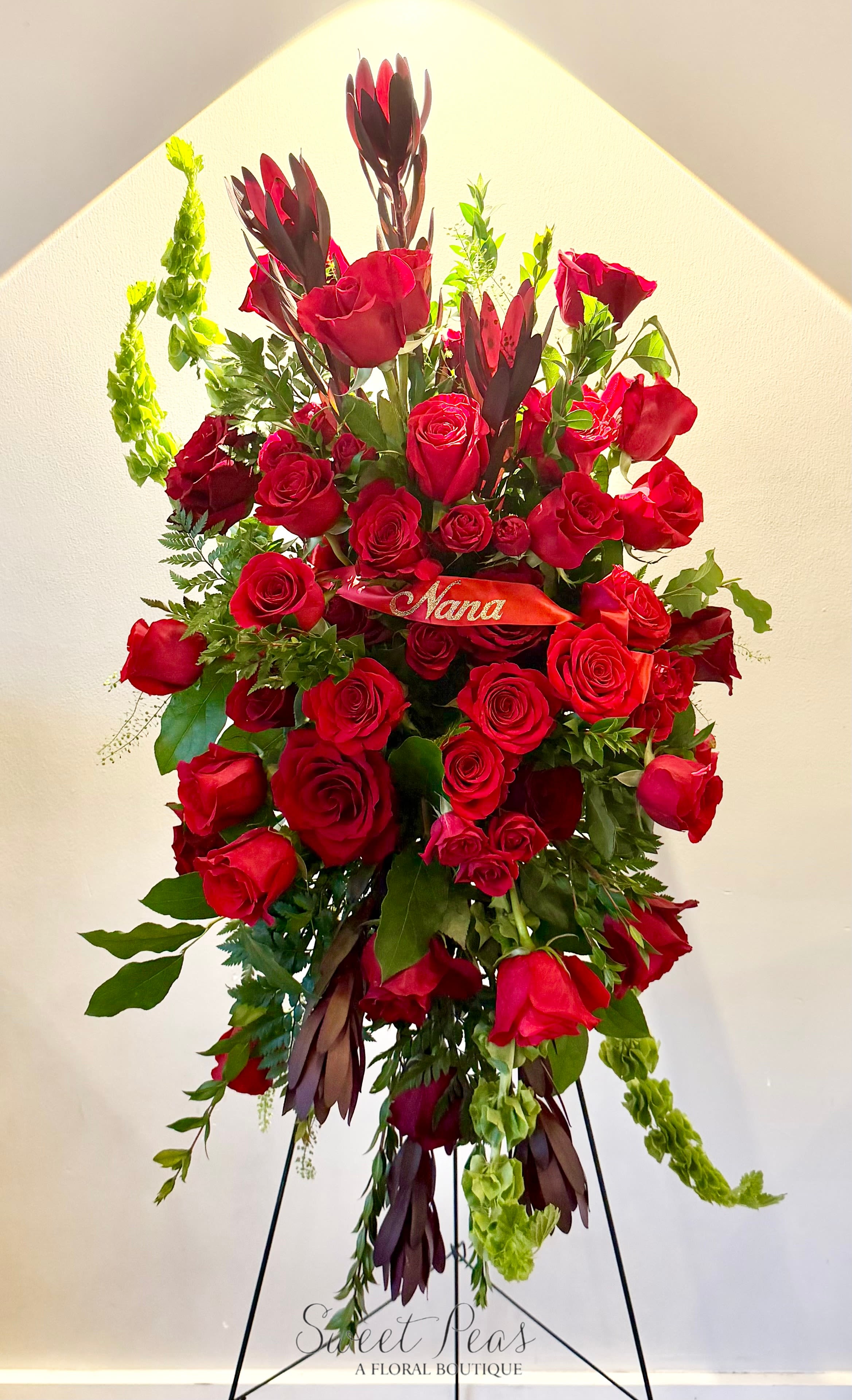 Crimson Remembrance Standing Spray - Striking and deeply heartfelt, Crimson Remembrance is a standing spray designed to honor and celebrate a life filled with love. Featuring an abundant display of rich red roses accented with lush greenery and textural elements, this arrangement symbolizes deep love, respect, and remembrance. Displayed on an easel and finished with a personalized ribbon, it creates a beautiful and meaningful tribute for funeral or memorial services.