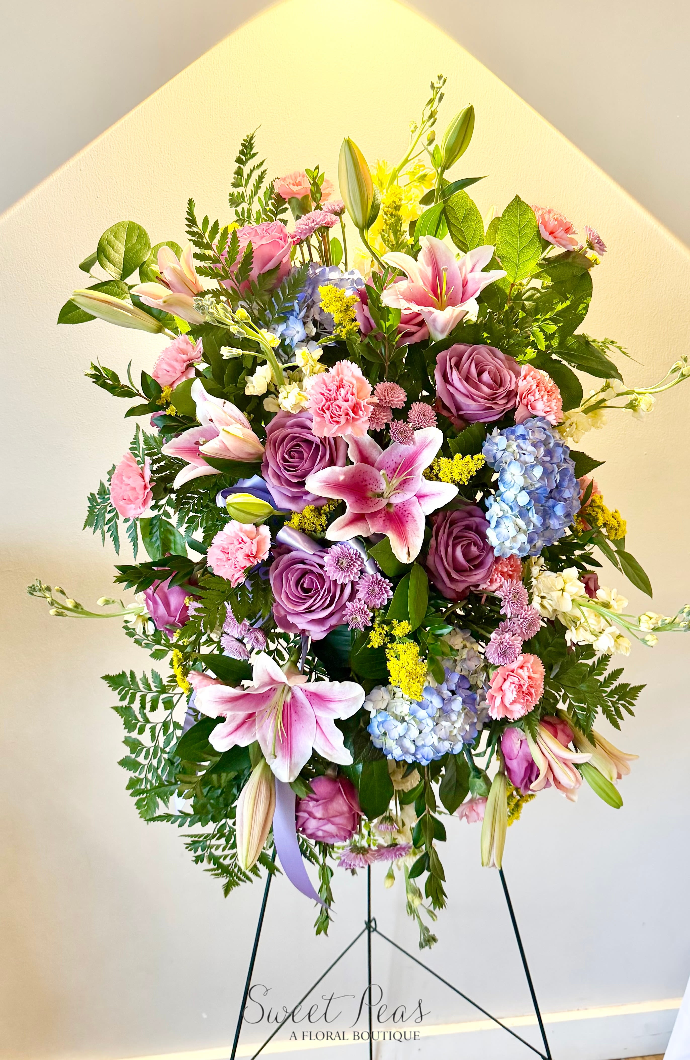 Heavenly Garden Tribute - Soft and uplifting, Heavenly Garden Tribute is a beautiful standing spray designed to celebrate a life with grace and love. Featuring a delicate blend of pink lilies, lavender roses, hydrangea, and soft seasonal blooms, this arrangement is accented with fresh greenery for a natural, garden-inspired feel. Displayed on an easel, it offers a gentle expression of sympathy, peace, and remembrance—bringing comfort while honoring a life beautifully lived.