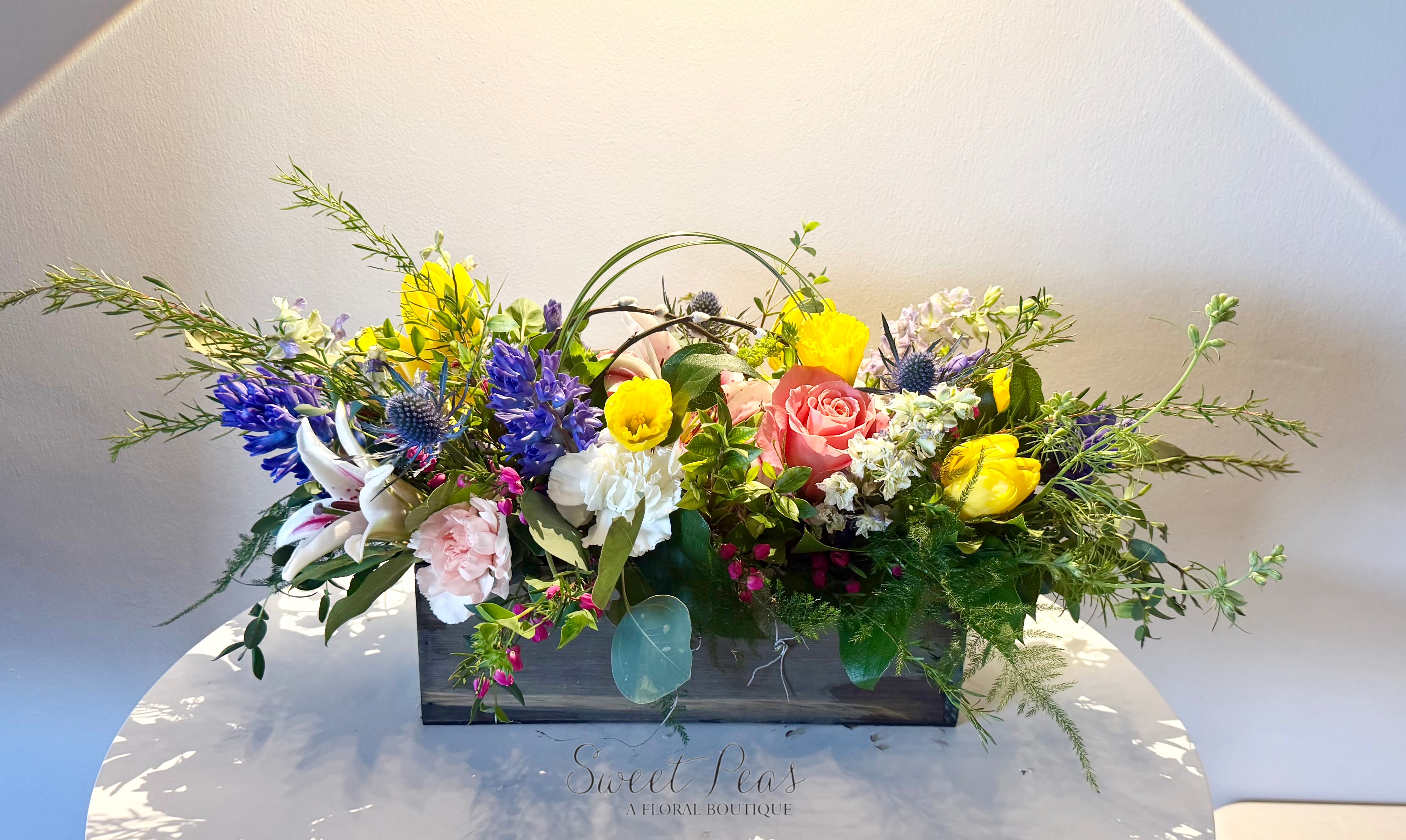 Easter Garden Gathering - Fresh and full of springtime joy, Easter Garden Gathering is a vibrant design inspired by the beauty of the season. Featuring a cheerful mix of tulips, daffodils, hyacinth, roses, and other seasonal blooms, this arrangement is filled with soft pastels and sunny pops of color. Designed in a rustic wooden box with airy greenery and natural movement, it captures the feeling of a blooming spring garden. Perfect for Easter celebrations or welcoming the new season, this piece brings warmth, renewal, and happiness to any space.