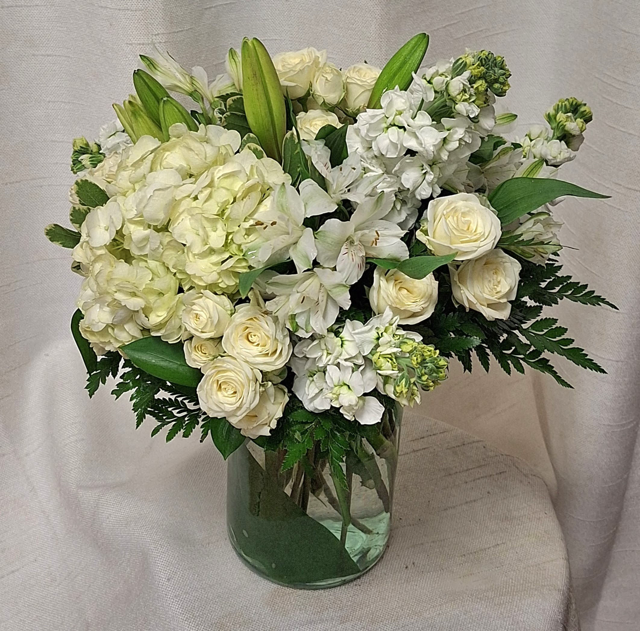 White and cream roses, hydrangeas, lilies, and stock arranged in a clear glass cylinder vase