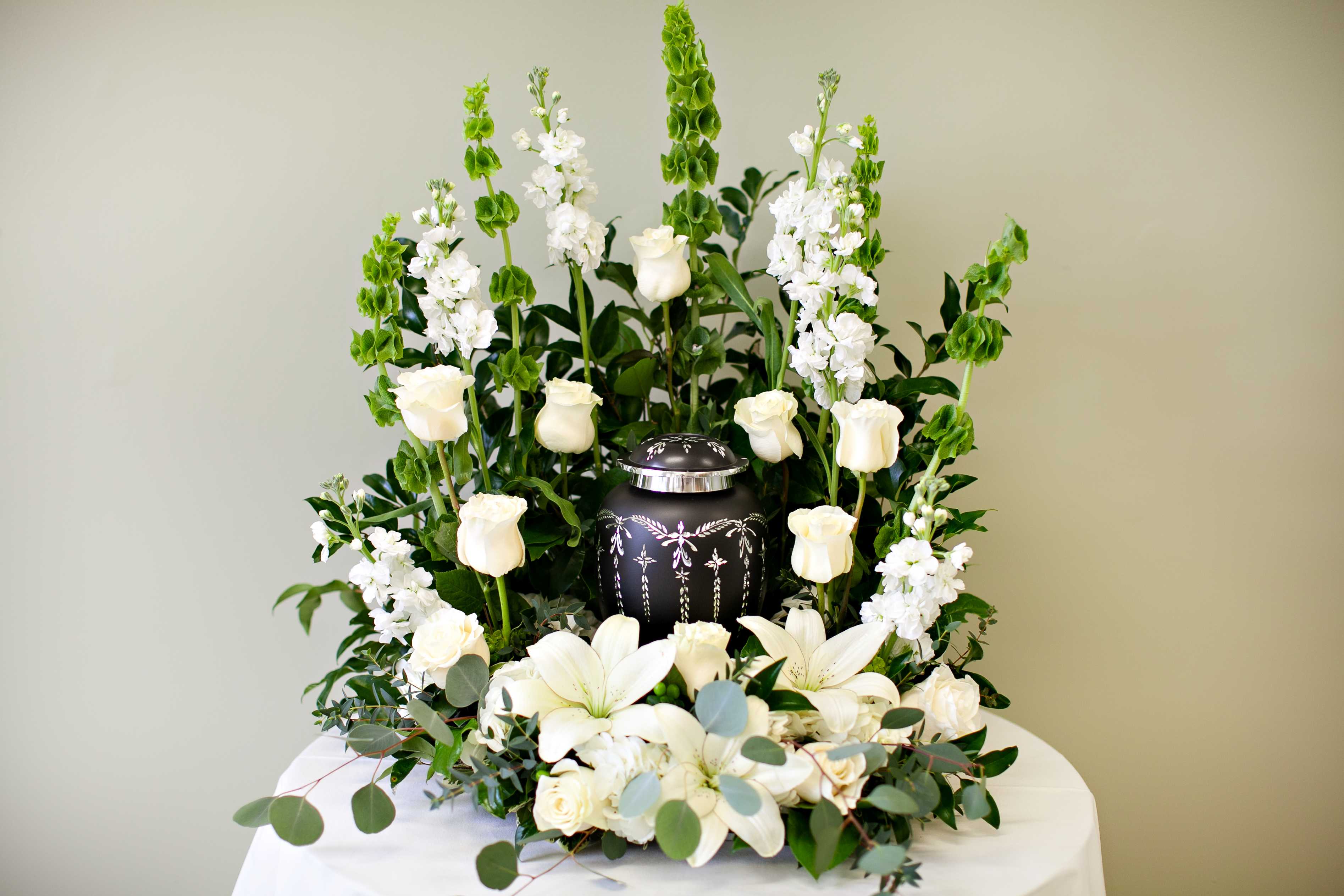 White roses and lilies surrounding a decorative urn in a tall white sympathy arrangement