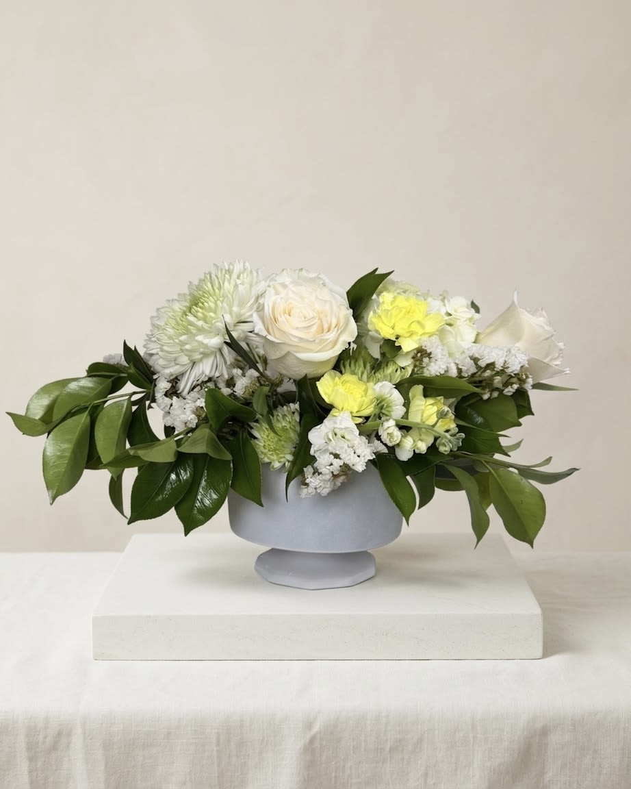 Citrine Whisper - A refined study in softness and light, Citrine Whisper blends creamy ivory blooms with delicate notes of pale yellow for a composition that feels both fresh and quietly luminous.  Centered around premium white roses and layered seasonal blooms, this arrangement is designed with intentional negative space and natural movement—allowing each stem to breathe and unfold gracefully. Subtle touches of soft yellow add warmth without overpowering, while polished greenery provides structure and contrast.  Designed in a modern vessel, this piece feels elevated yet approachable—perfect for sending a gesture that is thoughtful, clean, and effortlessly sophisticated. 