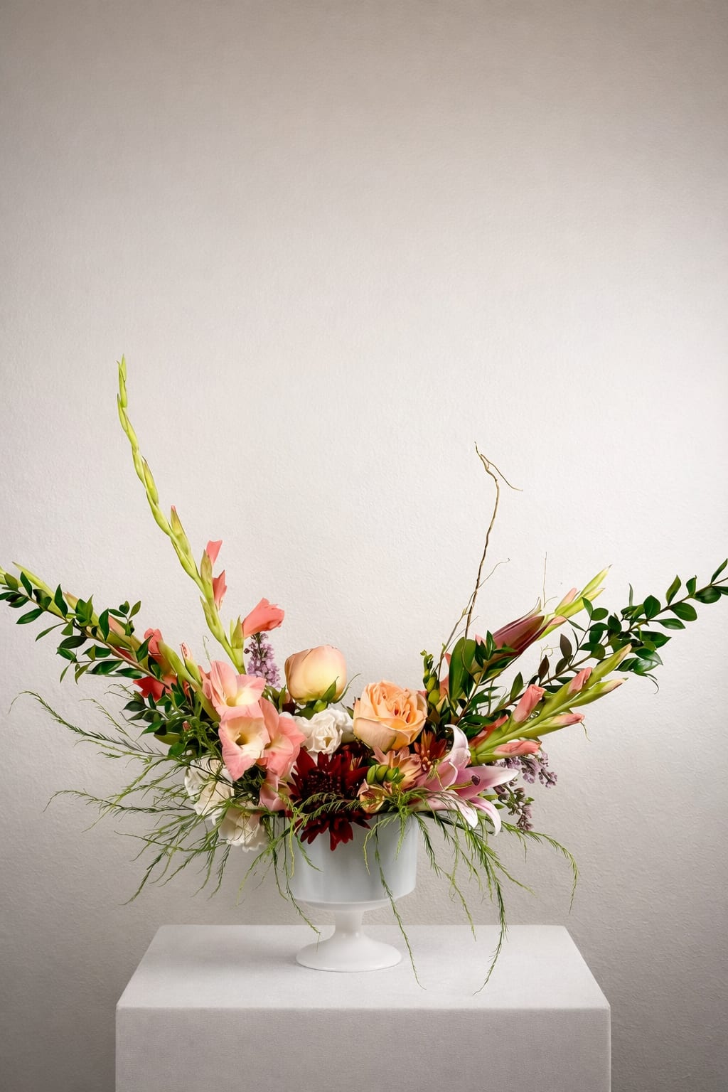 Coral Crescendo - The palette is soft yet vibrant, blending sun-washed peach, blush, and coral tones with fresh green elements for a look that feels modern, organic, and elevated. Designed in a clean white pedestal vessel, this arrangement balances structure and spontaneity—perfect for statement moments, intimate gatherings, or refined everyday luxury. 