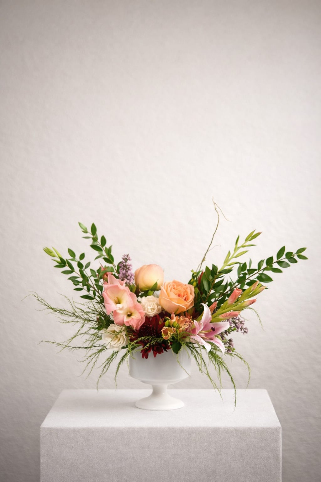 Coral Meadow  - With the vertical drama softened, this design feels more grounded and lush—featuring layered peach and coral roses, delicate lilies, and airy accent blooms nestled into abundant, flowing greenery.  The absence of taller spiking elements allows the composition to feel fuller and more intimate, while trailing greens and sculptural foliage create a relaxed, effortless silhouette. Designed in a classic white pedestal vessel, this piece evokes the feeling of a modern wild garden—refined, yet beautifully untamed.  Perfect for coffee tables, intimate dinners, or thoughtful gifting, Coral Meadow brings warmth and natural elegance into any space.