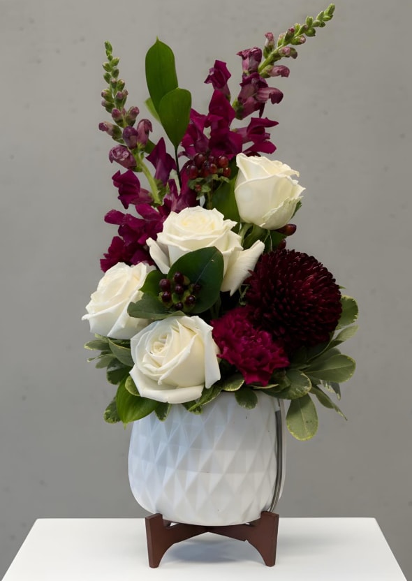 Crimson Elegance Vase - A rich and sophisticated arrangement featuring classic white roses paired with deep burgundy blooms, including snapdragons, carnations, and a statement mum. Accented with lush greenery and subtle berry detail, this design is arranged in a modern textured ceramic vase on a wooden stand—perfect for adding a touch of elegance to any space or sending a refined, heartfelt gesture.