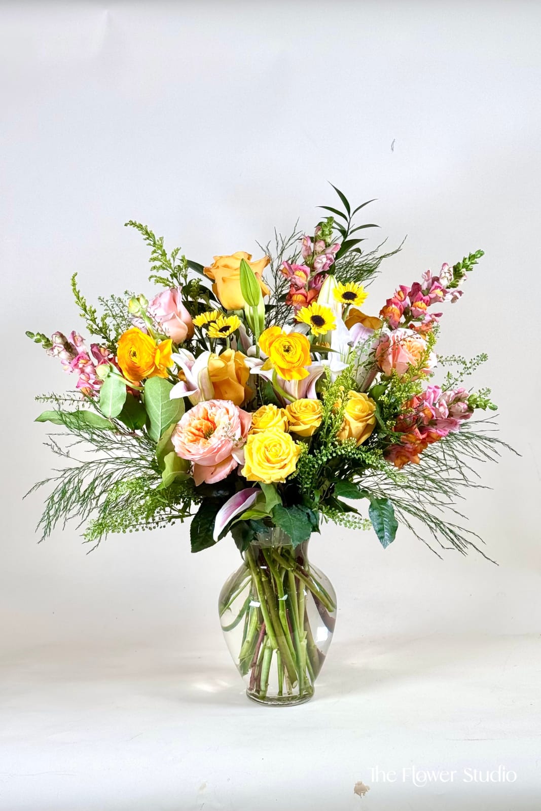 A lot of Spring - A bright and uplifting garden-style arrangement bursting with warm, sun-kissed tones of yellow, peach, and soft pink. Featuring a cheerful mix of roses, ranunculus, lilies, and delicate accent blooms, this design is layered with airy greenery for a natural, organic feel. Arranged in a clear glass vase, the stems add to its fresh-from-the-garden charm, making it perfect for celebrations, thank-yous, or simply bringing a little sunshine into any space. Size: H-22' W-20'