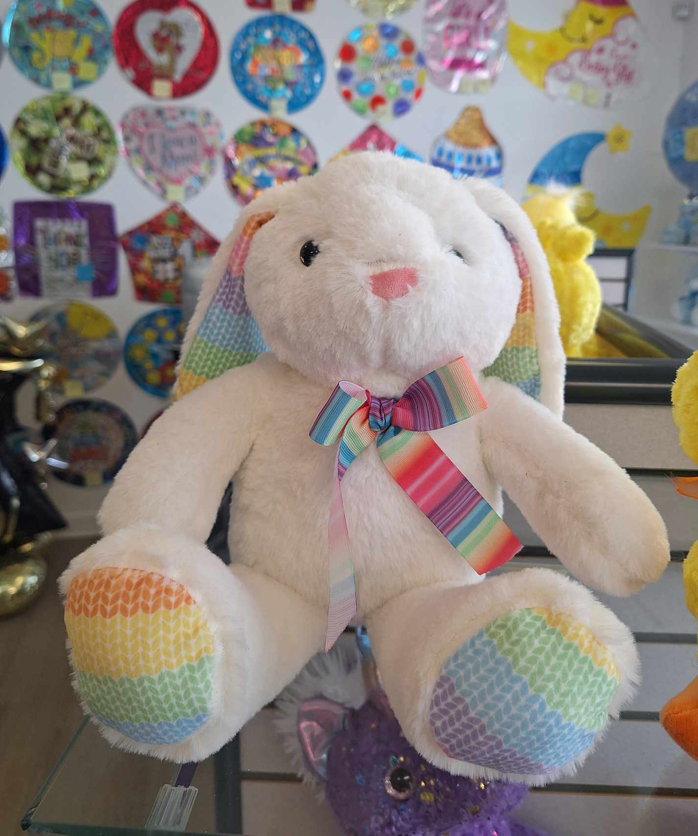 White plush bunny with rainbow ears, paws, and bow sitting in a gift shop display.