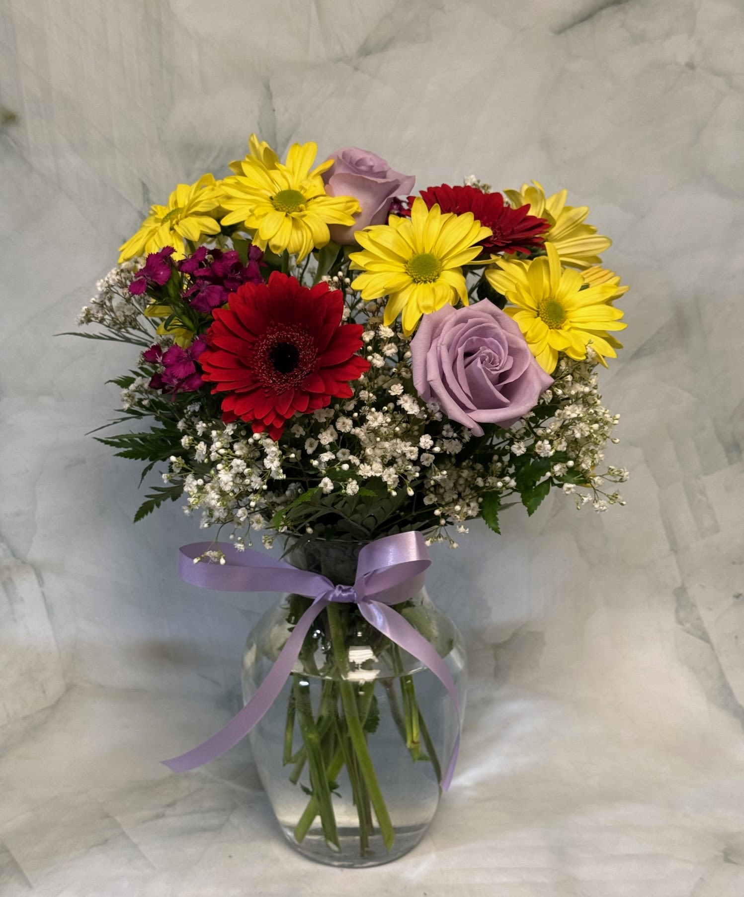 Mixed bouquet of yellow daisies, red gerbera daisies and lavender roses in a clear vase with a purple ribbon