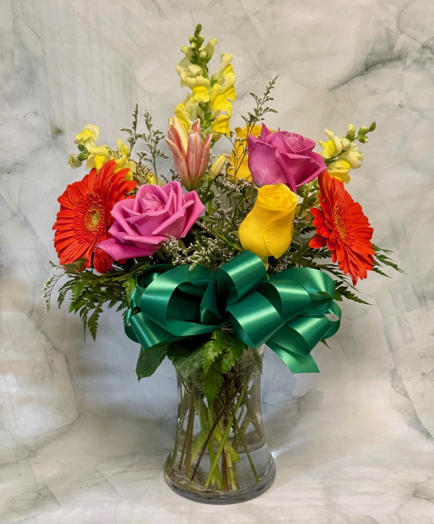 Mixed bouquet of pink, yellow, and orange flowers in a clear vase with a large green ribbon bow