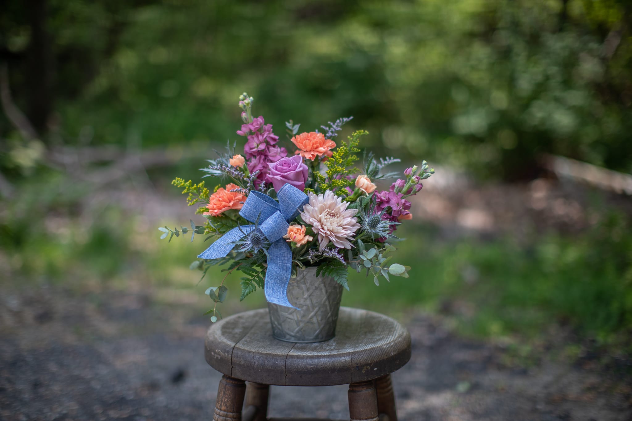 Mixed arrangement of peach carnations, lavender rose, and pink mum in a tin pot with a blue bow.