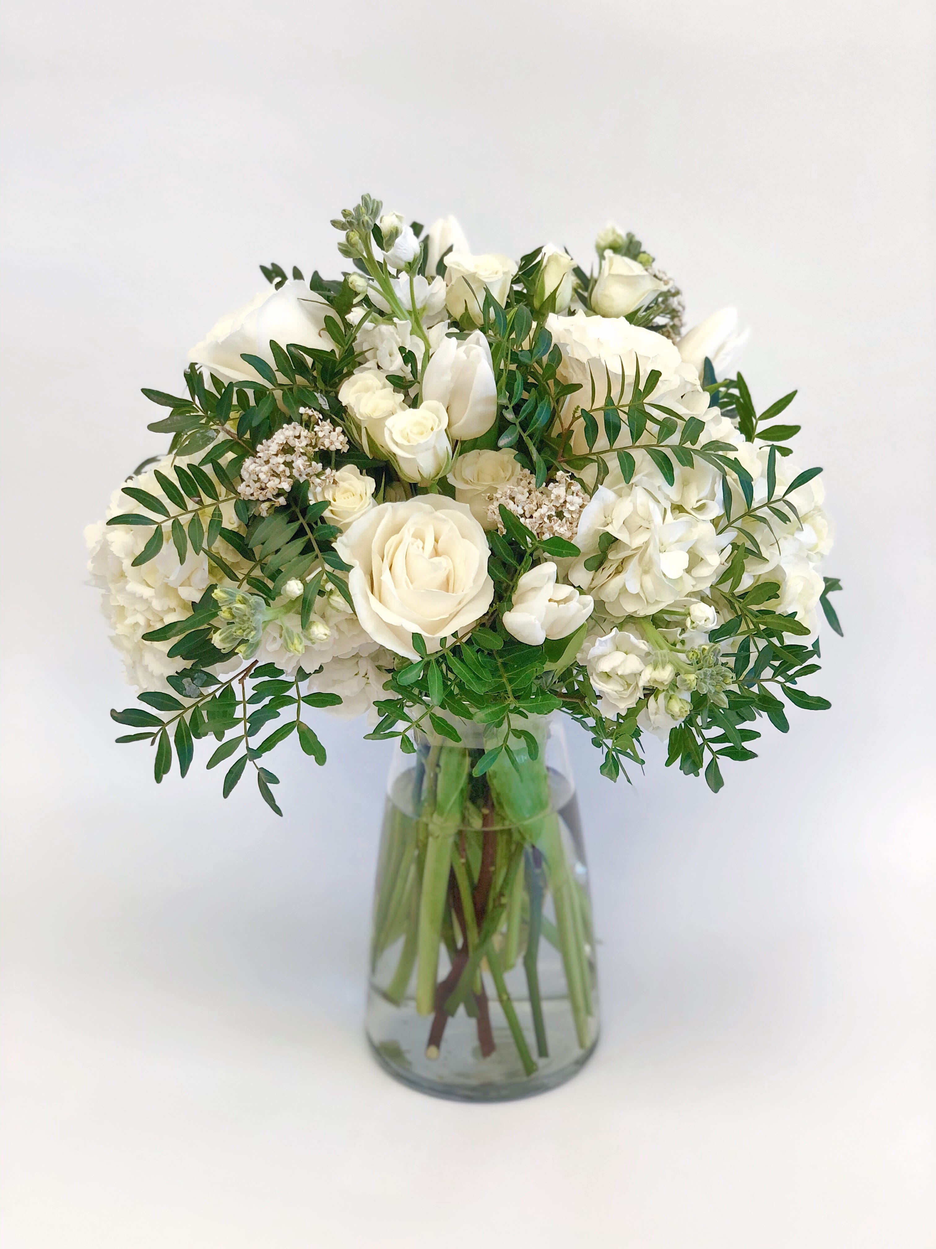 Vase Arrangement White and Green - This elegant vase arrangement features a stunning mix of fresh, seasonal flowers artfully designed to bring warmth and beauty to any setting. Each bouquet is thoughtfully composed with a variety of vibrant blooms, such as roses, stock, hydrangeas, spray roses, and greenery, creating a rich tapestry of color, texture, and fragrance.
