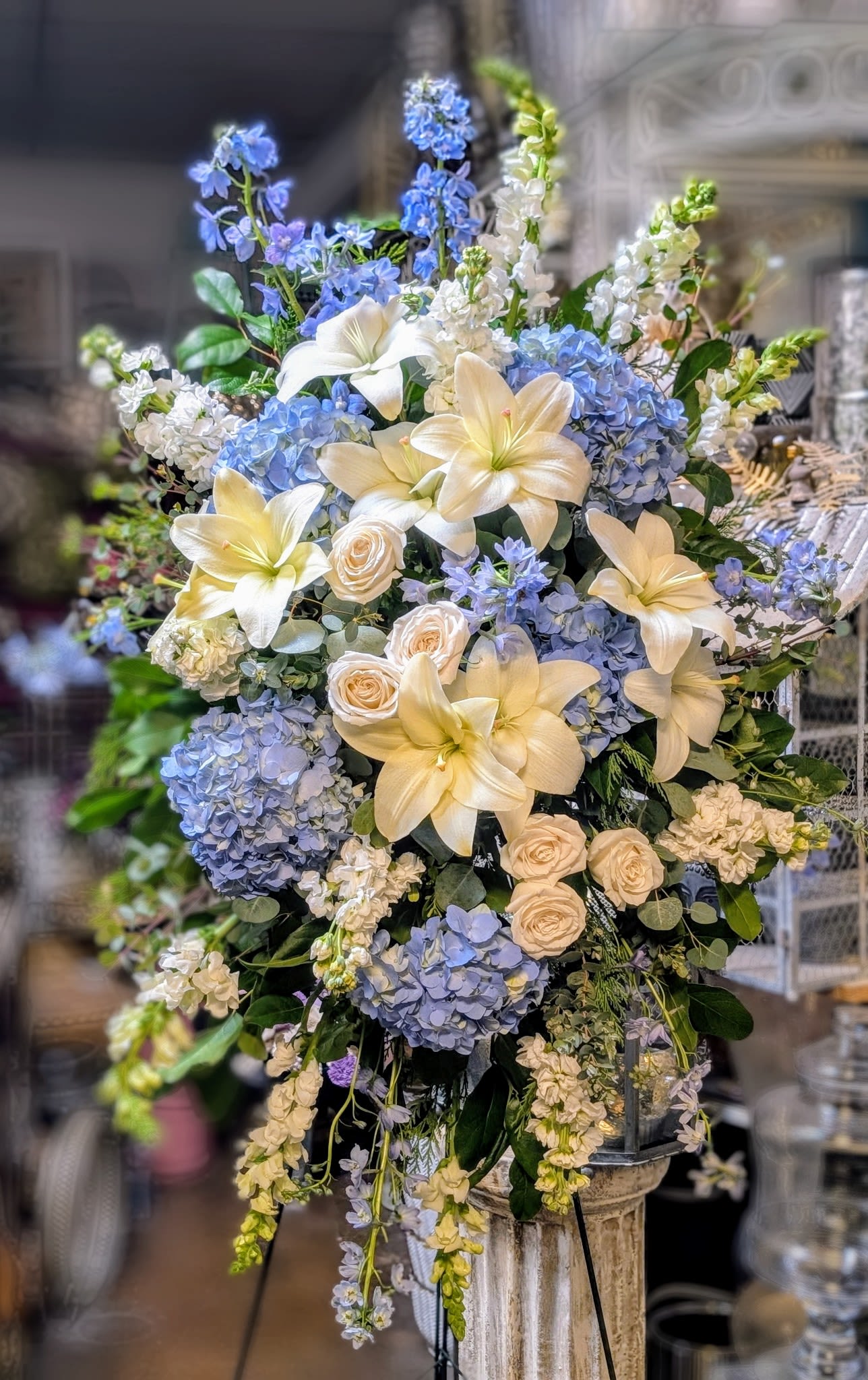 Tall arrangement of white lilies, cream roses, and blue hydrangeas on a pedestal