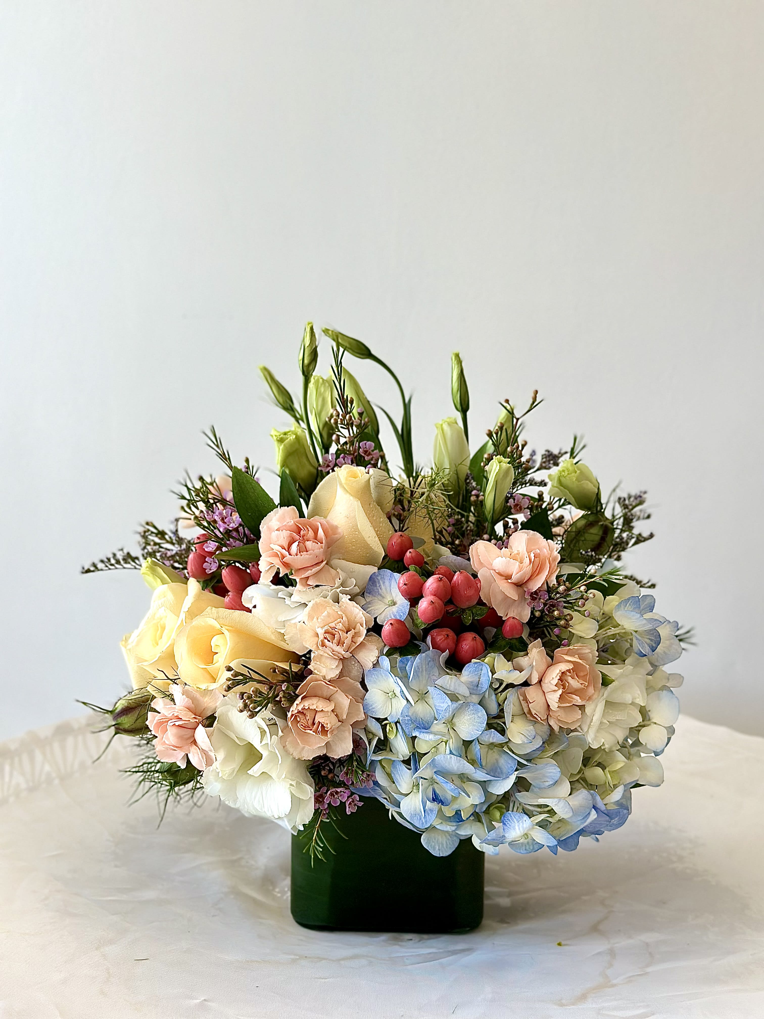 Low mixed arrangement of peach carnations, yellow roses and blue hydrangeas in a dark green glass cube vase