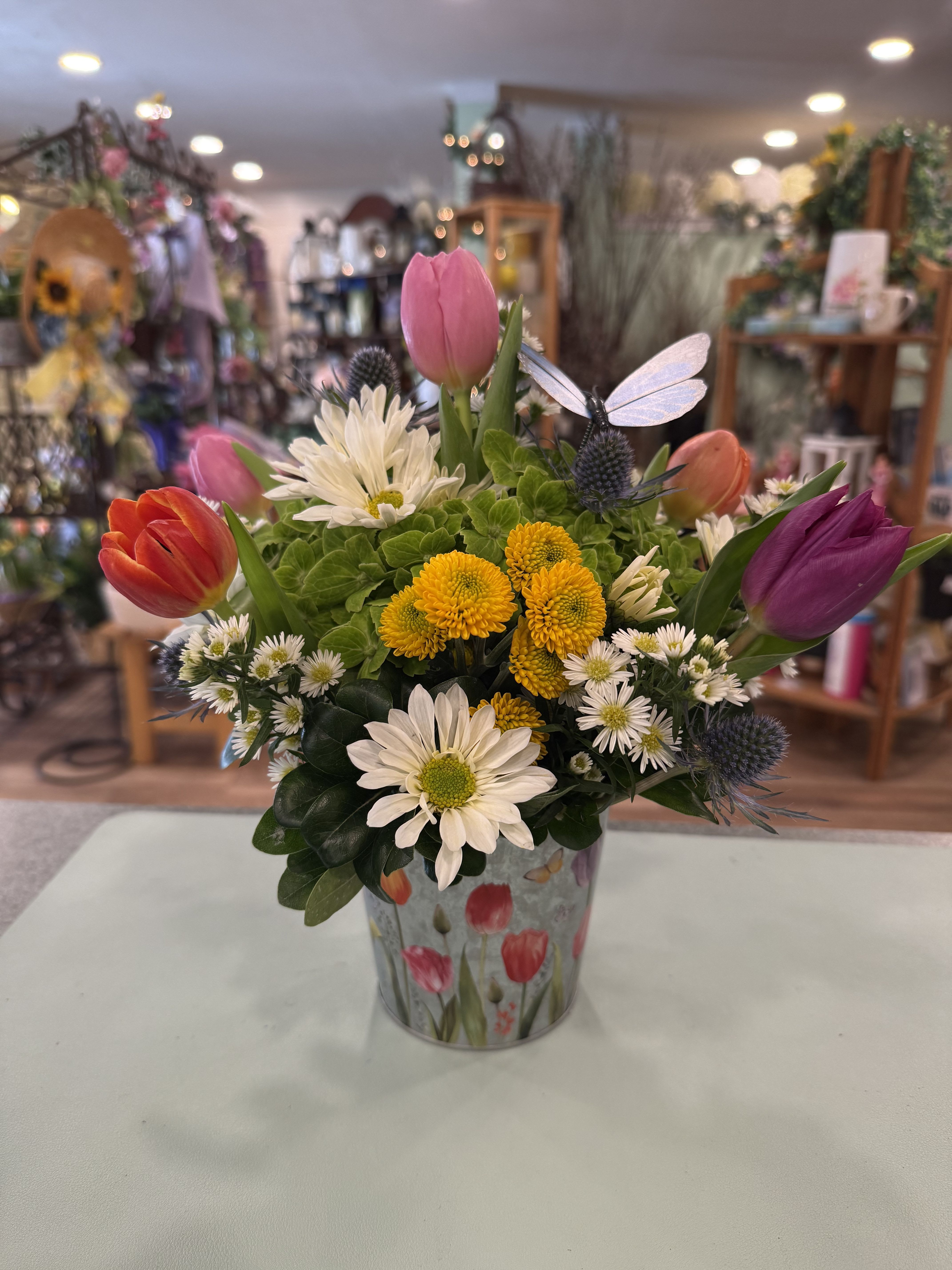 Tulip Garden (TUG) - Tulips, thistle, yellow buttons, white daisies and hydrangea make up this colorful spring arrangement designed in a tulip printed galvanized pot. Approx. 12 inches tall.  ***Colors and flower type may vary. 