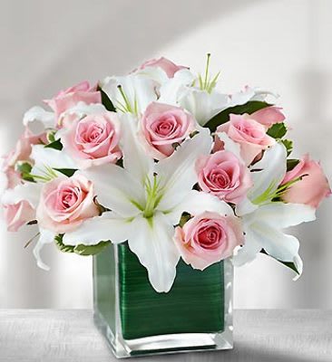 Pink roses and white lilies in a clear glass cube vase lined with a green leaf.