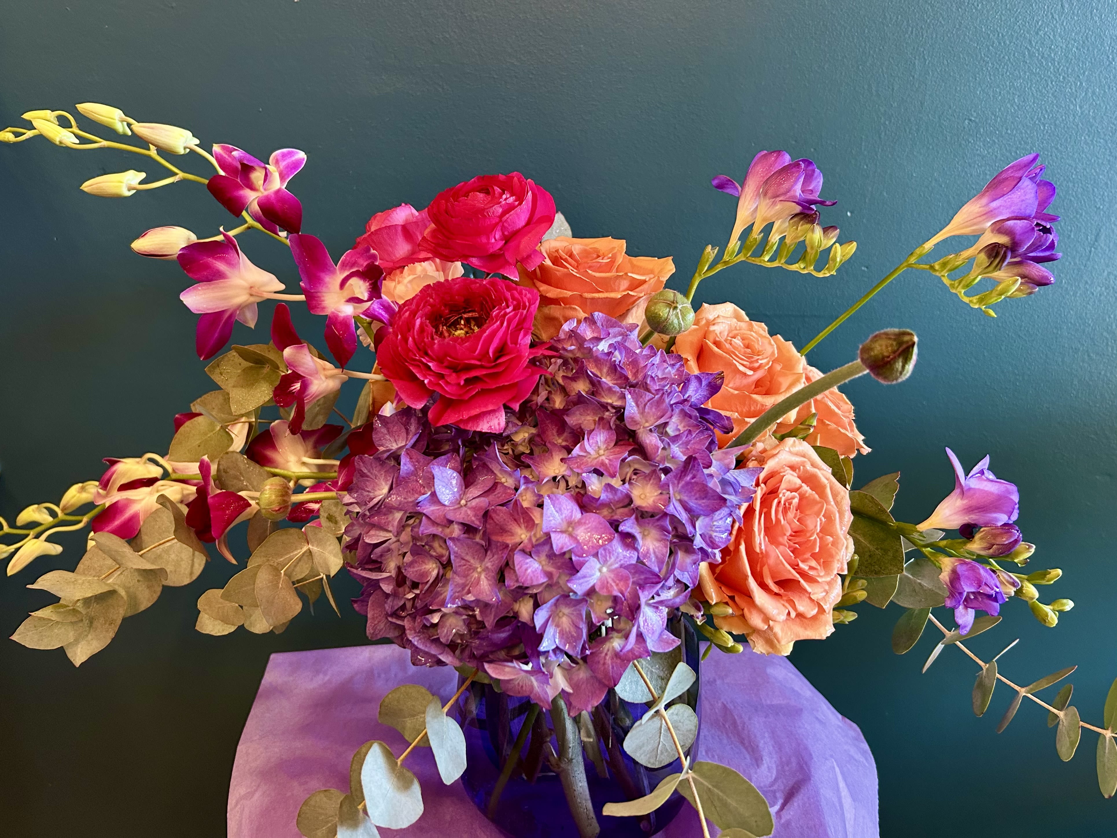Botanical Beauty - A lush design that will wow any flower lover! Overflowing with beautiful seasonal blooms, at their best and freshest. A bold color palette of purples, magentas and melon will bring joy to anyone who receives this thoughtfully curated bouquet. 
