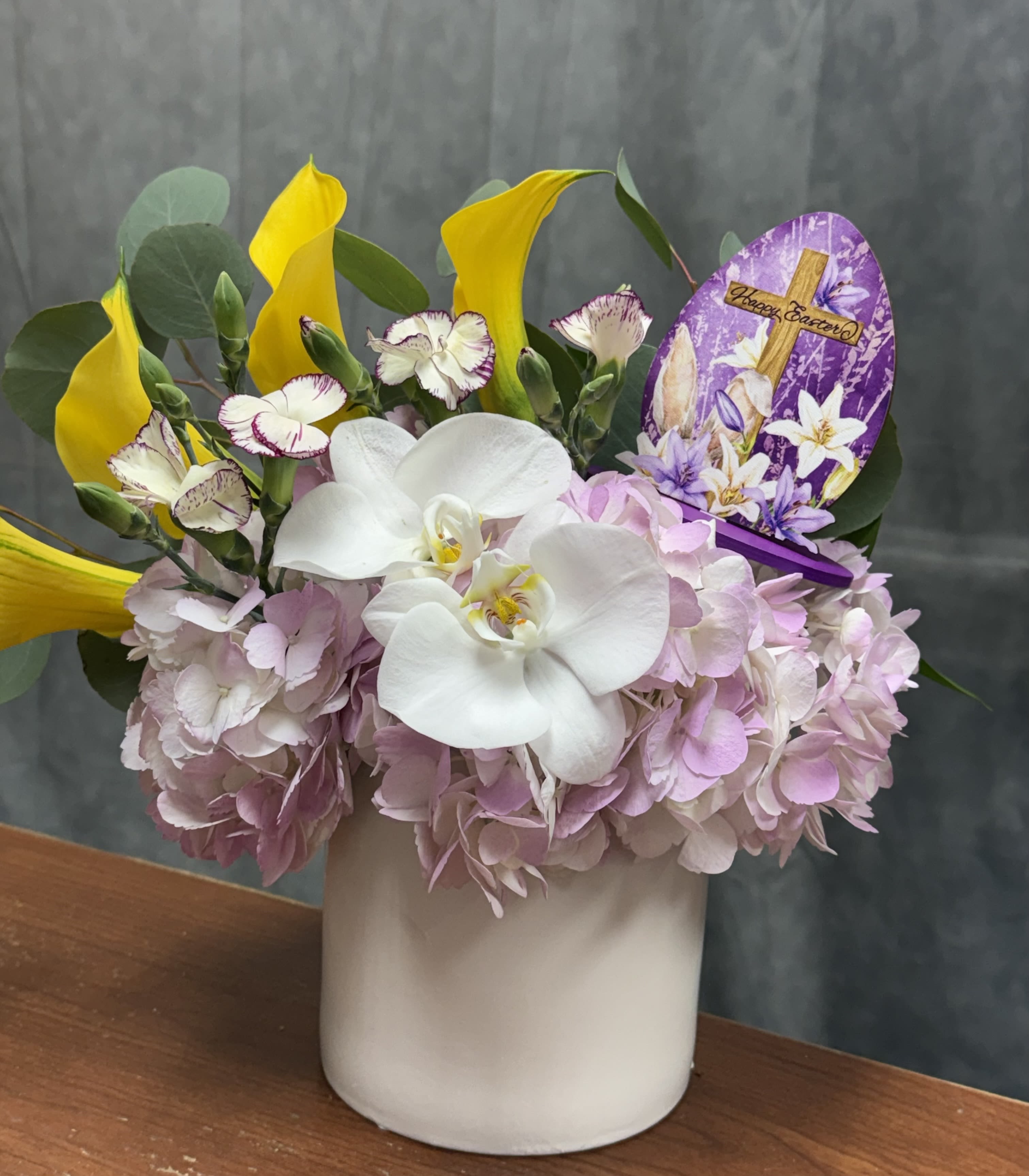 He is Risen Blooms  - A joyful mix of pastel blooms and vibrant yellow calla lilies, paired with a stunning white orchid symbolizing purity and renewal. Finished with an Easter cross keepsake, this arrangement is a heartfelt way to celebrate the season.