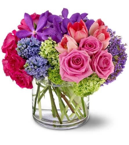 Colorful arrangement of pink roses, tulips, and mixed purple flowers in a clear glass vase