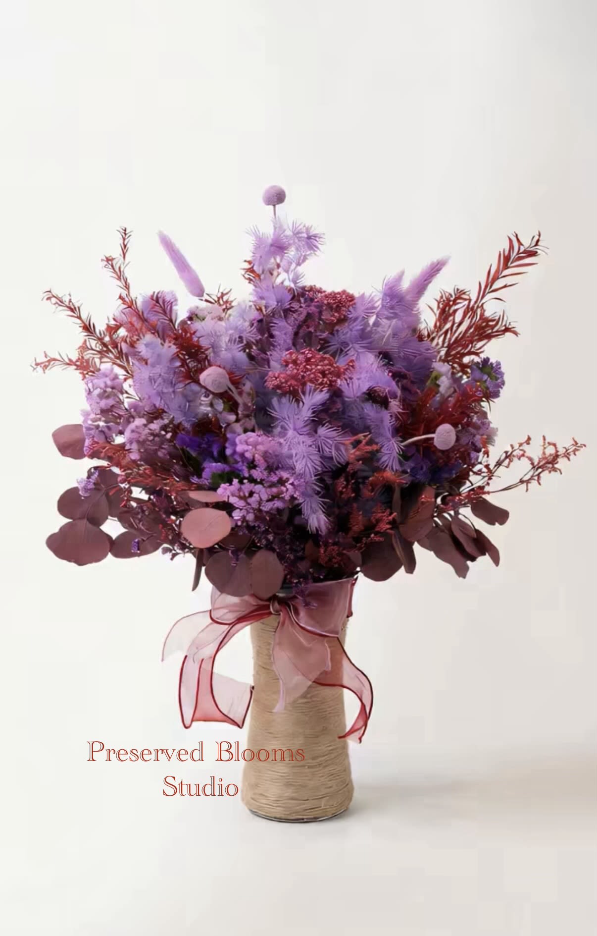 Tall bouquet of lavender and burgundy preserved flowers in a rope-wrapped vase with a pink ribbon bow