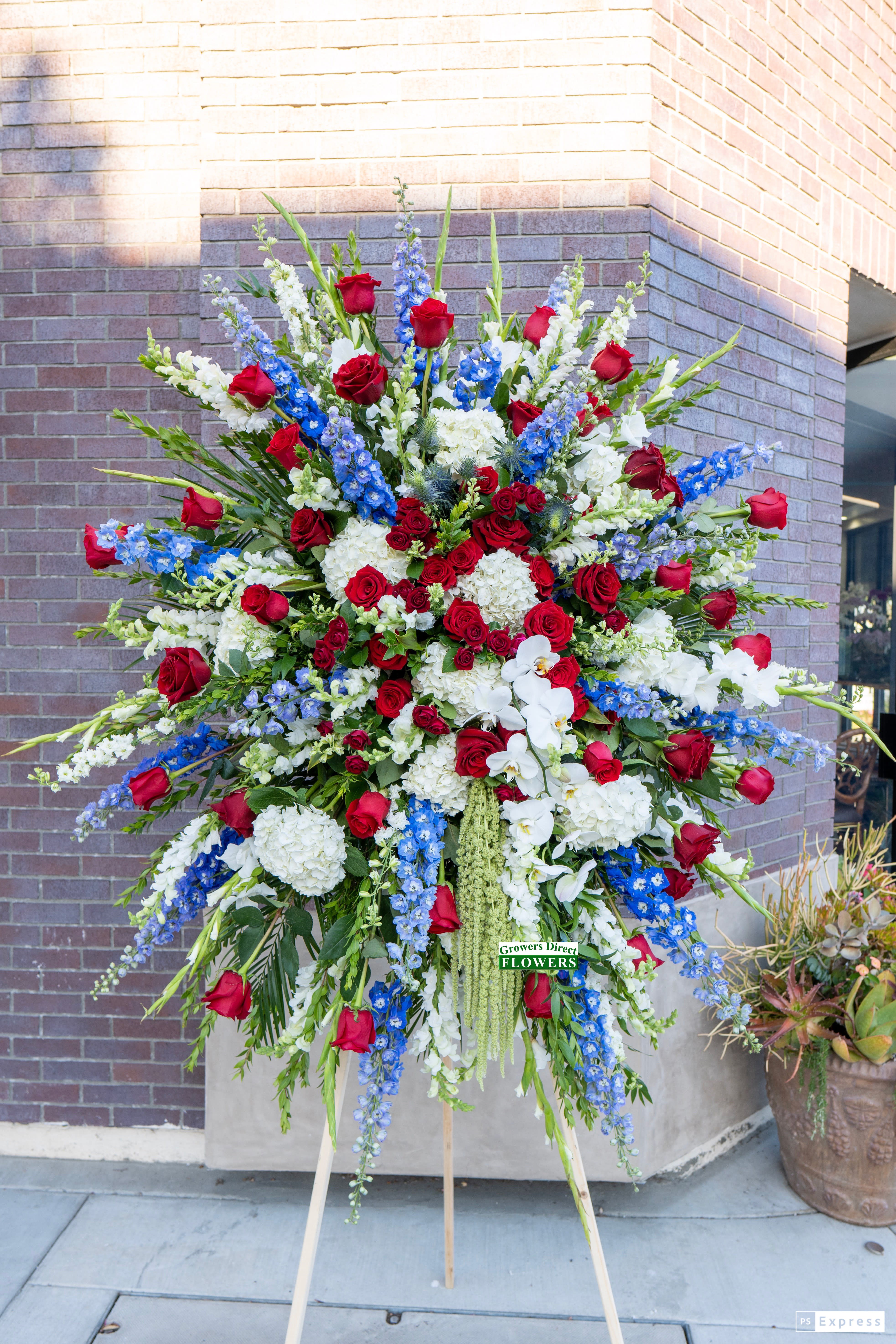 Tall standing spray with red roses, white hydrangeas and orchids, and blue delphinium on an easel