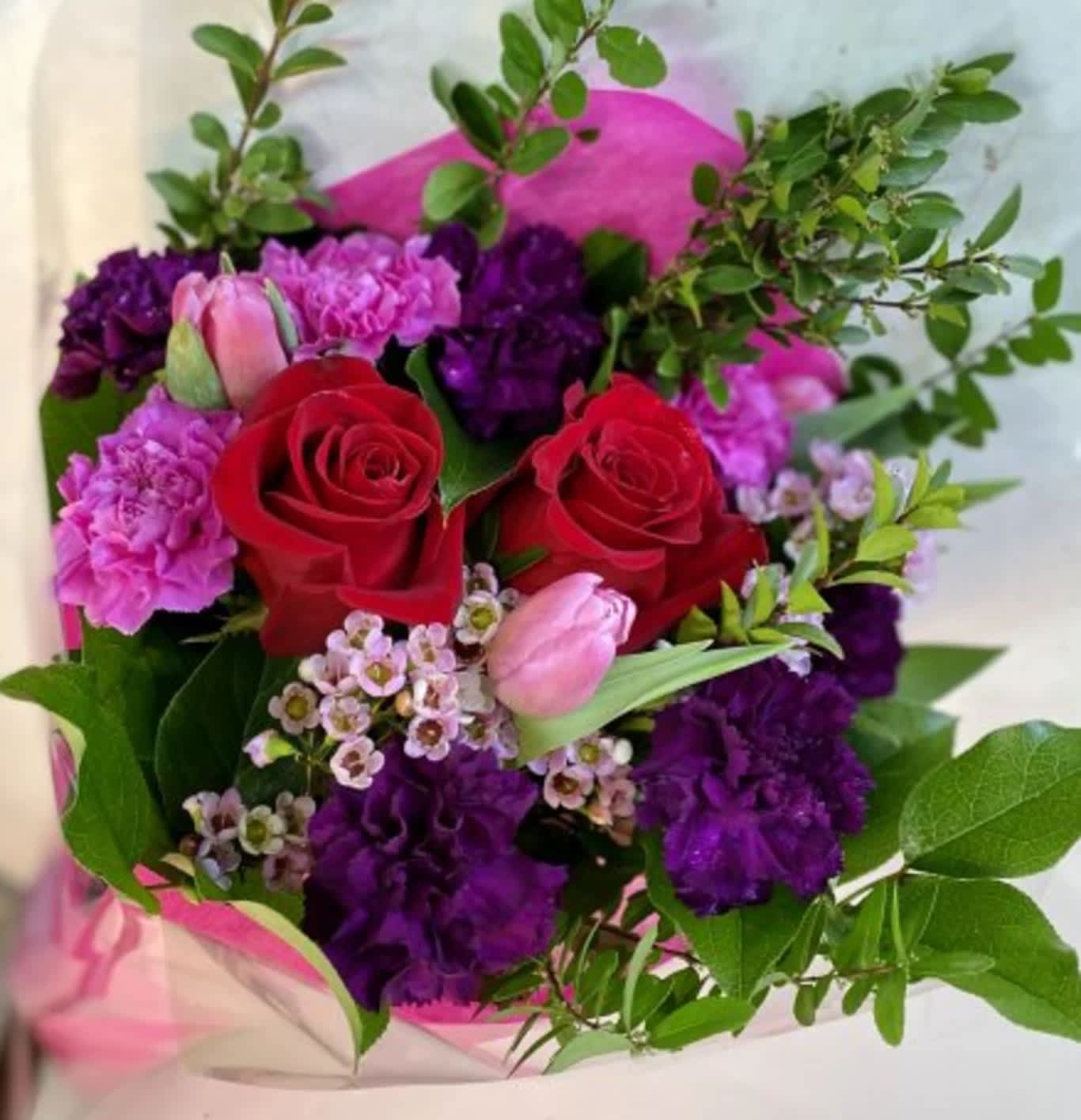 Hand-tied bouquet with red roses, purple carnations, pink tulips, and small white blossoms in pink wrap.