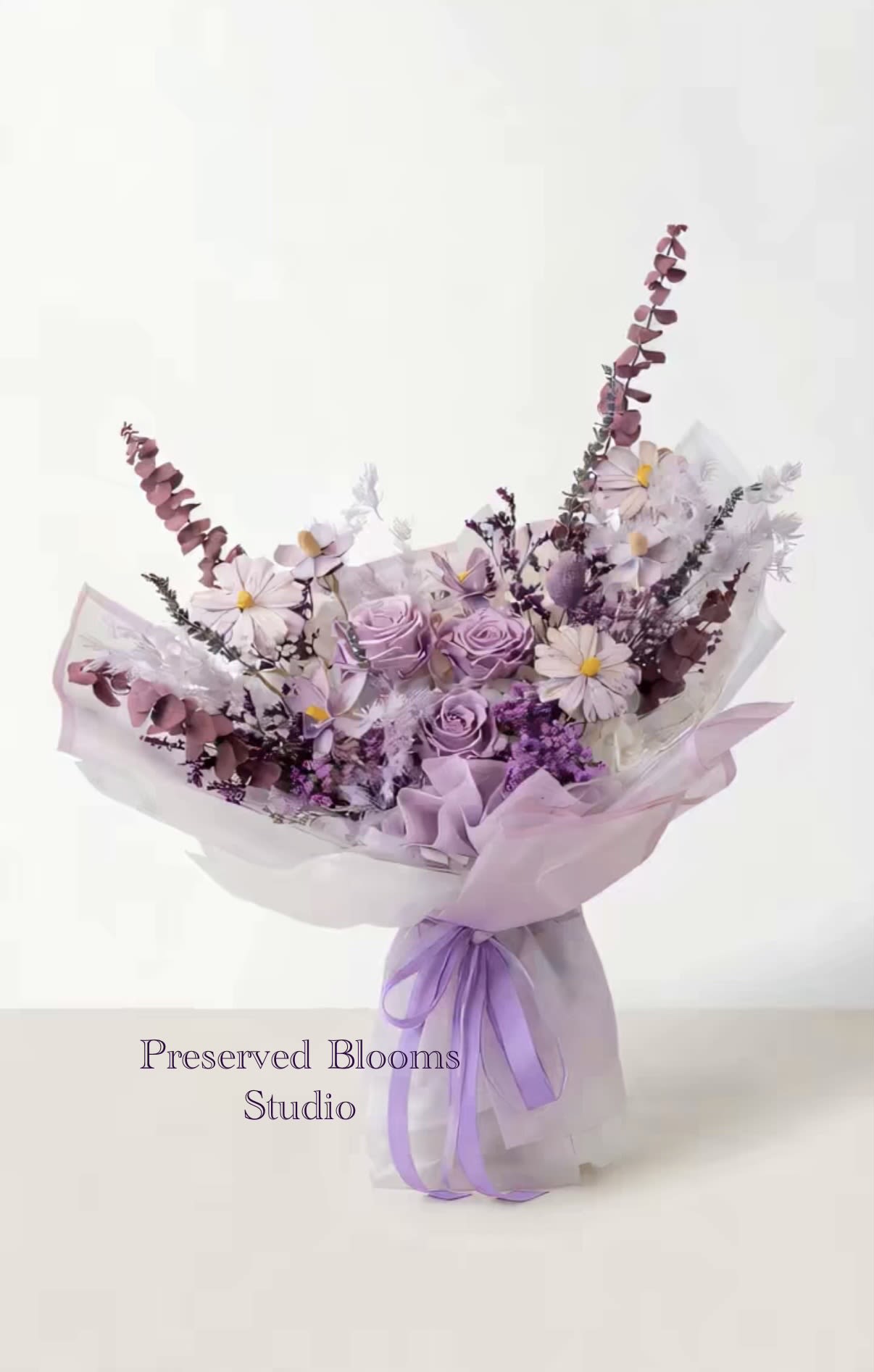 Lavender and white bouquet of roses and daisies wrapped in sheer paper with a purple ribbon bow