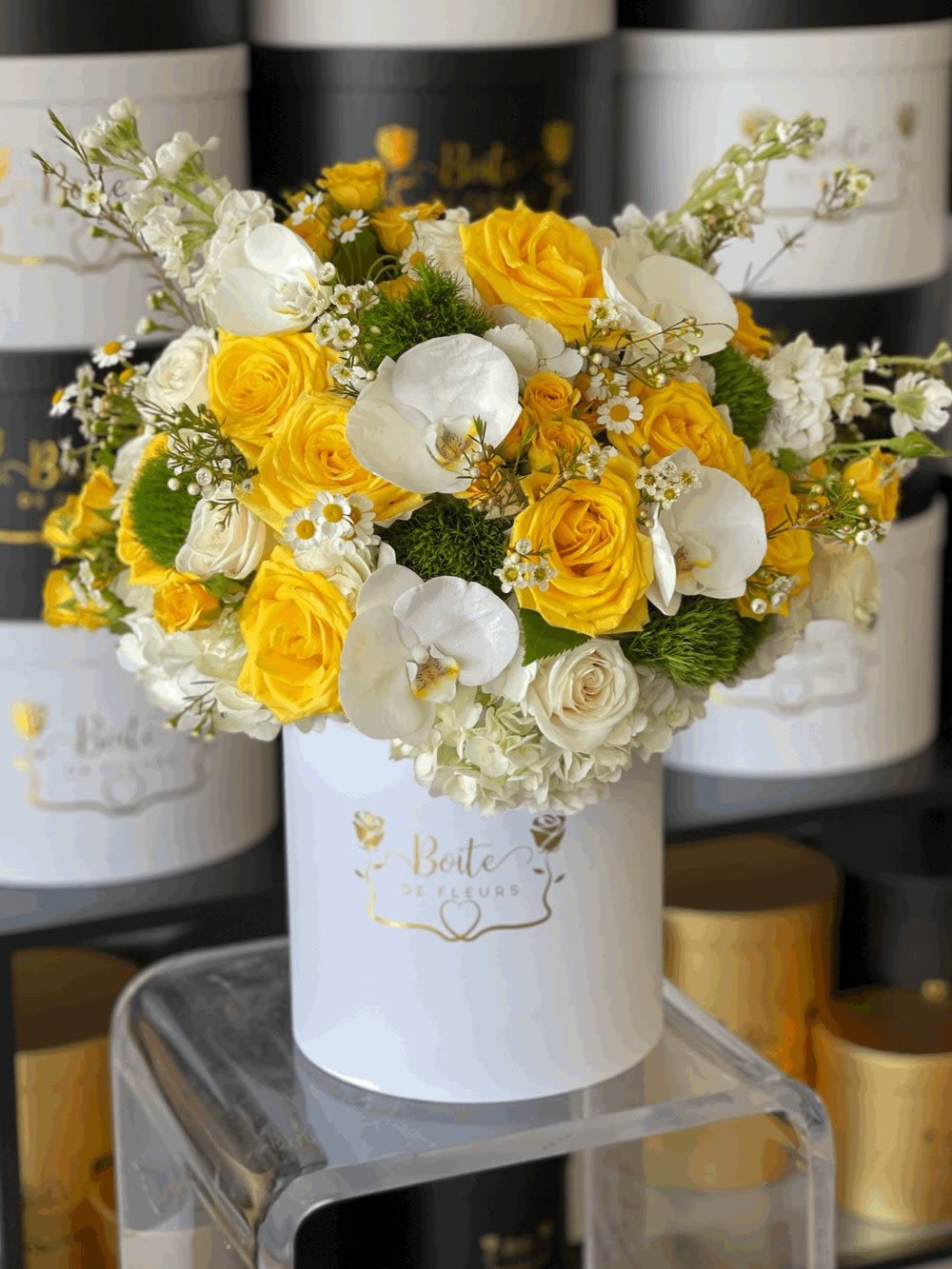 Round hatbox arrangement of yellow roses, white orchids, hydrangeas, and small daisies in a white box.