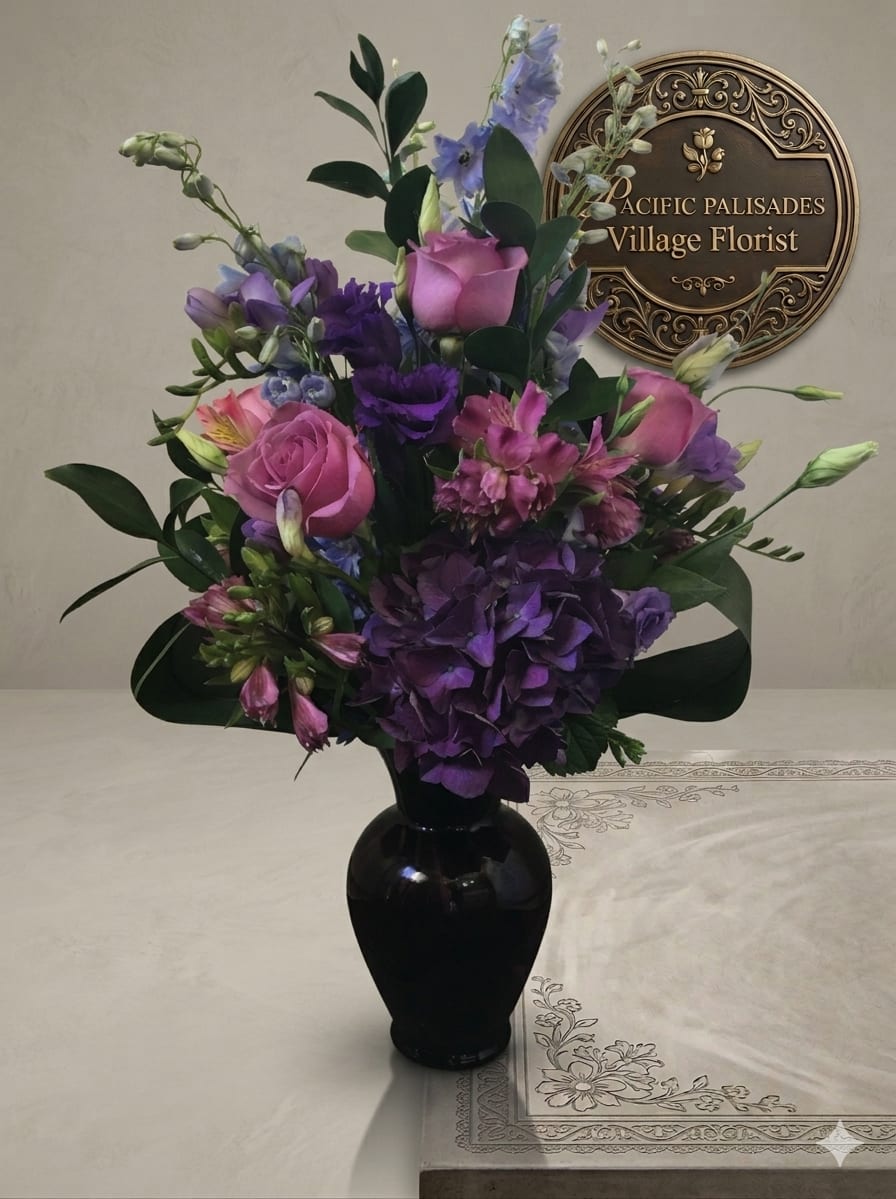 Visions of Violet - A rich, moody arrangement in shades of violet and blush, featuring soft pink roses layered with deep purple hydrangea and lisianthus, accented by alstroemeria and airy blue delphinium. The design feels romantic and dramatic, with lush textures and a sophisticated color palette set in a sleek dark vase.
