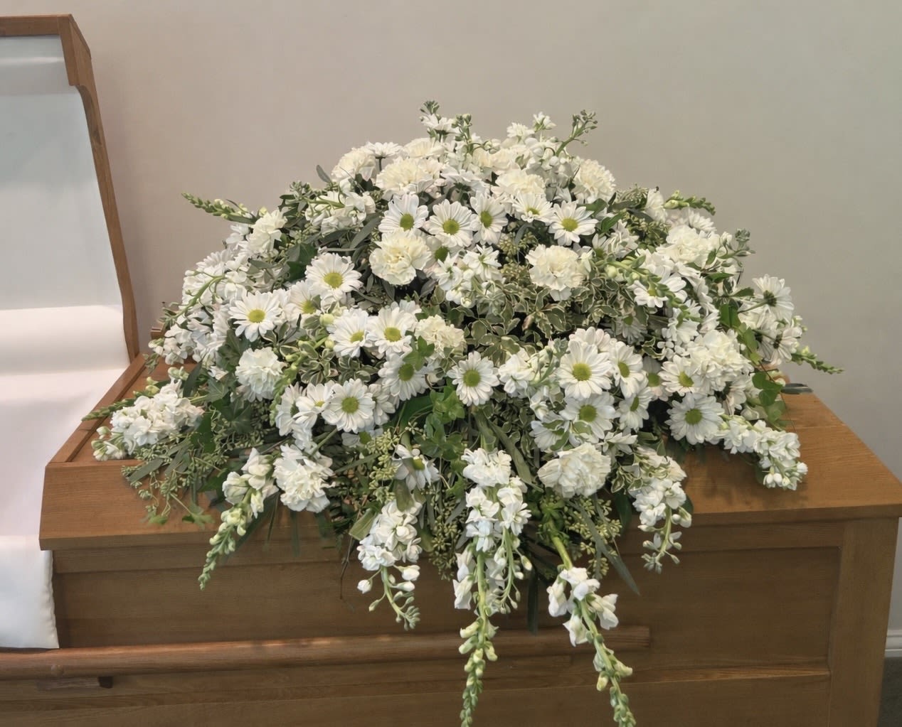 Ivory Field Casket Spray - Ivory Field  Ivory Field is a soft, natural-style casket spray designed with a full, flowing presence across the casket. White daisies, carnations, stock, and snapdragon are layered with textured greenery to create a design that feels open, airy, and quietly elegant.  The combination of rounded blooms and vertical line flowers gives this piece both fullness and gentle movement, allowing it to feel substantial without becoming heavy or overly structured. The all-white palette keeps the look clean and timeless, offering a sense of peace and simplicity that speaks for itself.  This design is ideal for families who want something classic, natural, and understated—an expression of love that feels calm, sincere, and beautifully composed.  Each arrangement is created with the freshest stems available. Subtle variations may occur while preserving the overall style, color palette, and design.  Available for casket flower delivery through Aspen Tower Florist, serving Greensburg and Shelbyville along with Rushville, Batesville, Westport, North Vernon, Oldenburg, St. Paul, Waldron, Milroy, and Hope. 
