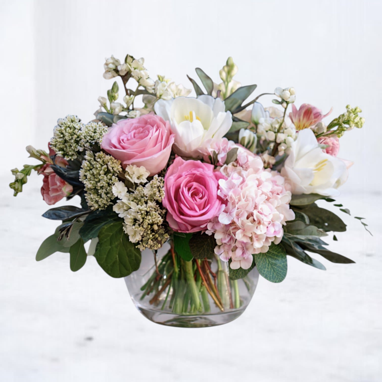 Low vase arrangement of pink roses, white blooms, and blush hydrangea in a clear glass bowl.
