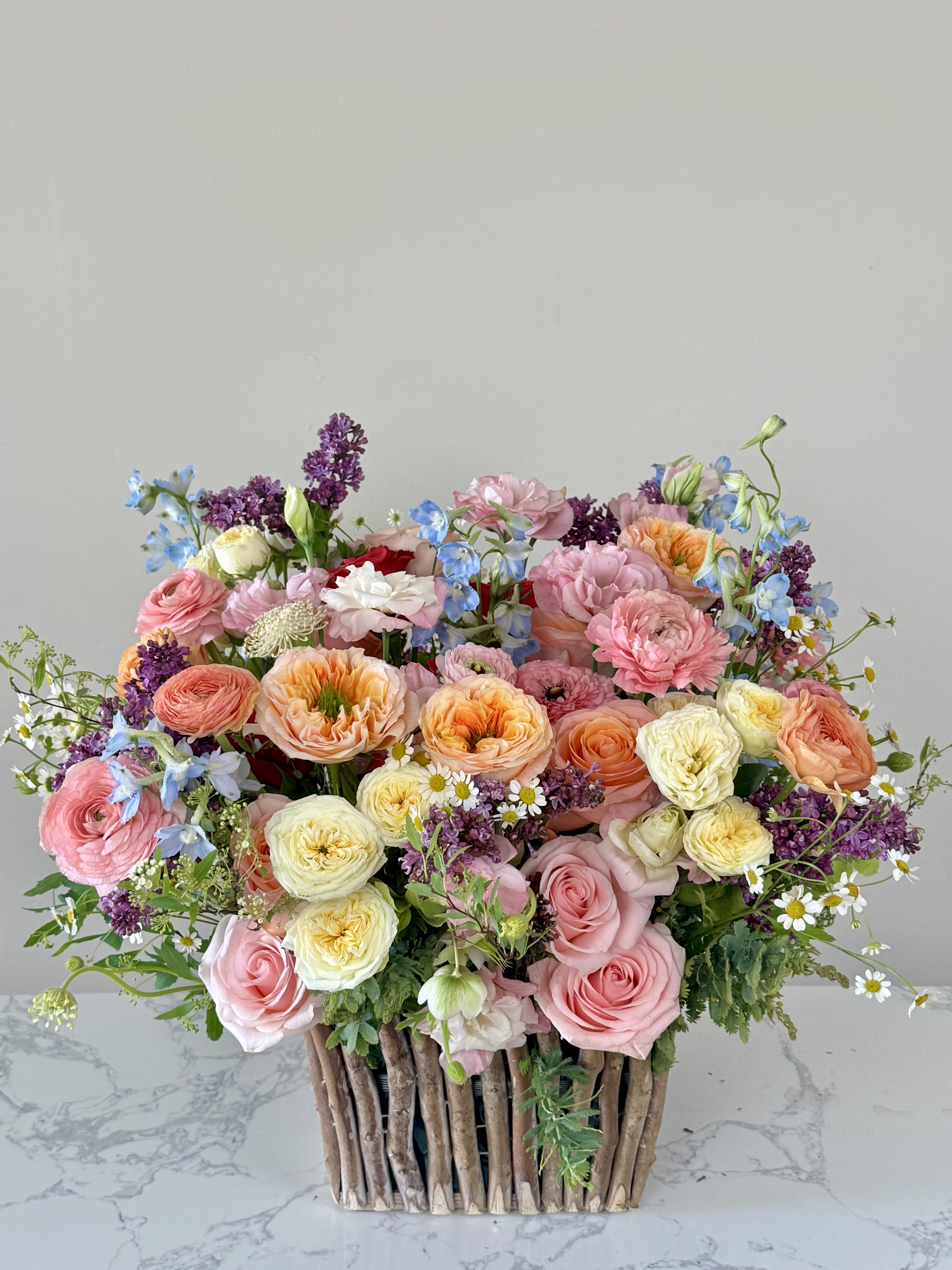 Easter Garden - Celebrate the beauty of spring with this lush, garden-inspired Easter arrangement bursting with soft pastels and cheerful blooms. Featuring delicate blush roses, buttery ranunculus, peach garden roses, and airy sprays of lilac and chamomile, this design captures the essence of a fresh spring morning. Accents of light blue delphinium and seasonal greenery add a whimsical, meadow-like feel, all thoughtfully arranged in a rustic wooden container.  Perfect for Easter gatherings, brunch tables, or as a heartfelt seasonal gift, this arrangement brings warmth, color, and a touch of natural elegance to any space.