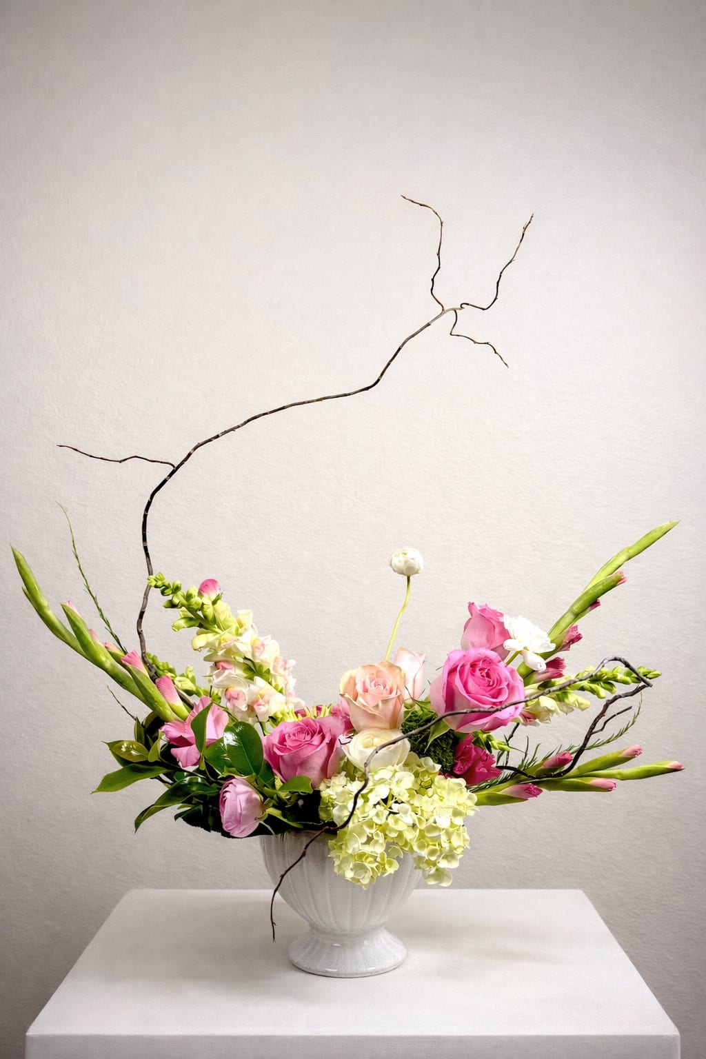 Spring Fling - A sculptural expression of softness and movement, Spring Fling blends romantic garden roses in shades of pink and blush with delicate hydrangea and airy snapdragons. Graceful branching adds an organic, architectural line, creating a sense of upward motion and quiet drama.  Designed in a classic white compote, this arrangement balances lush florals with negative space—offering a modern, gallery-like presence that feels both refined and effortlessly natural. Perfect as a statement centerpiece or an elevated gesture of elegance. 