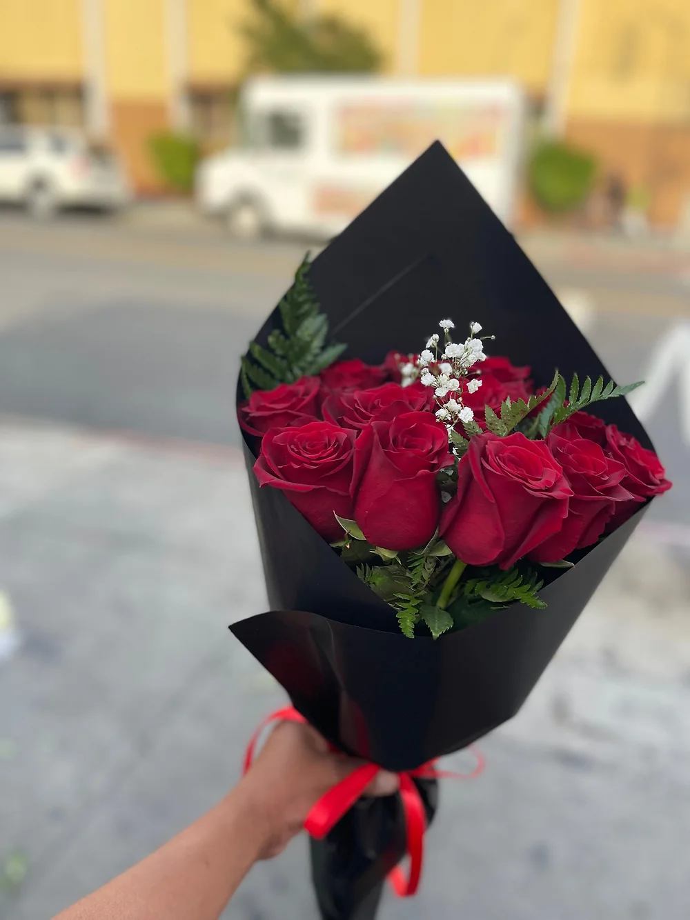 Handheld bouquet of red roses with white filler flowers wrapped in black paper and red ribbon