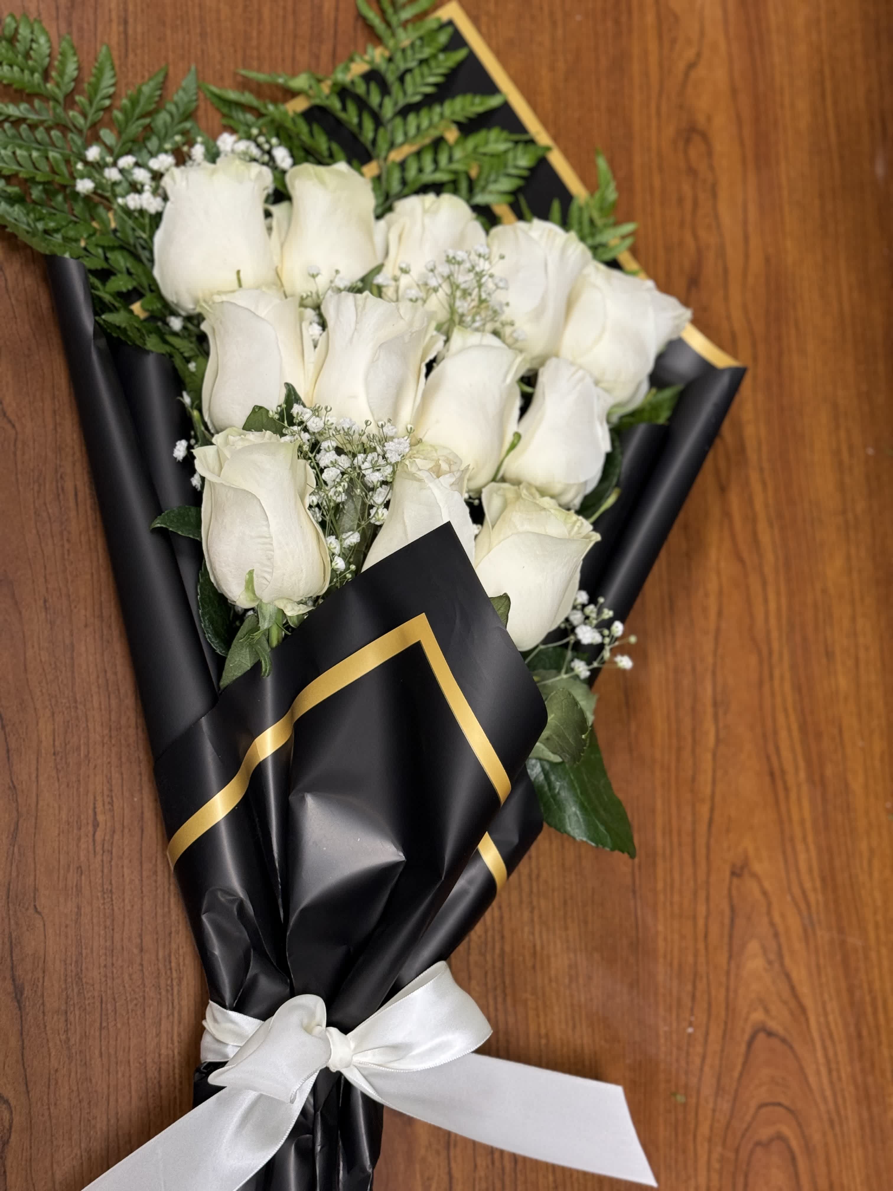 Wrapped bouquet of white roses with baby's breath in black paper with gold trim and white ribbon.
