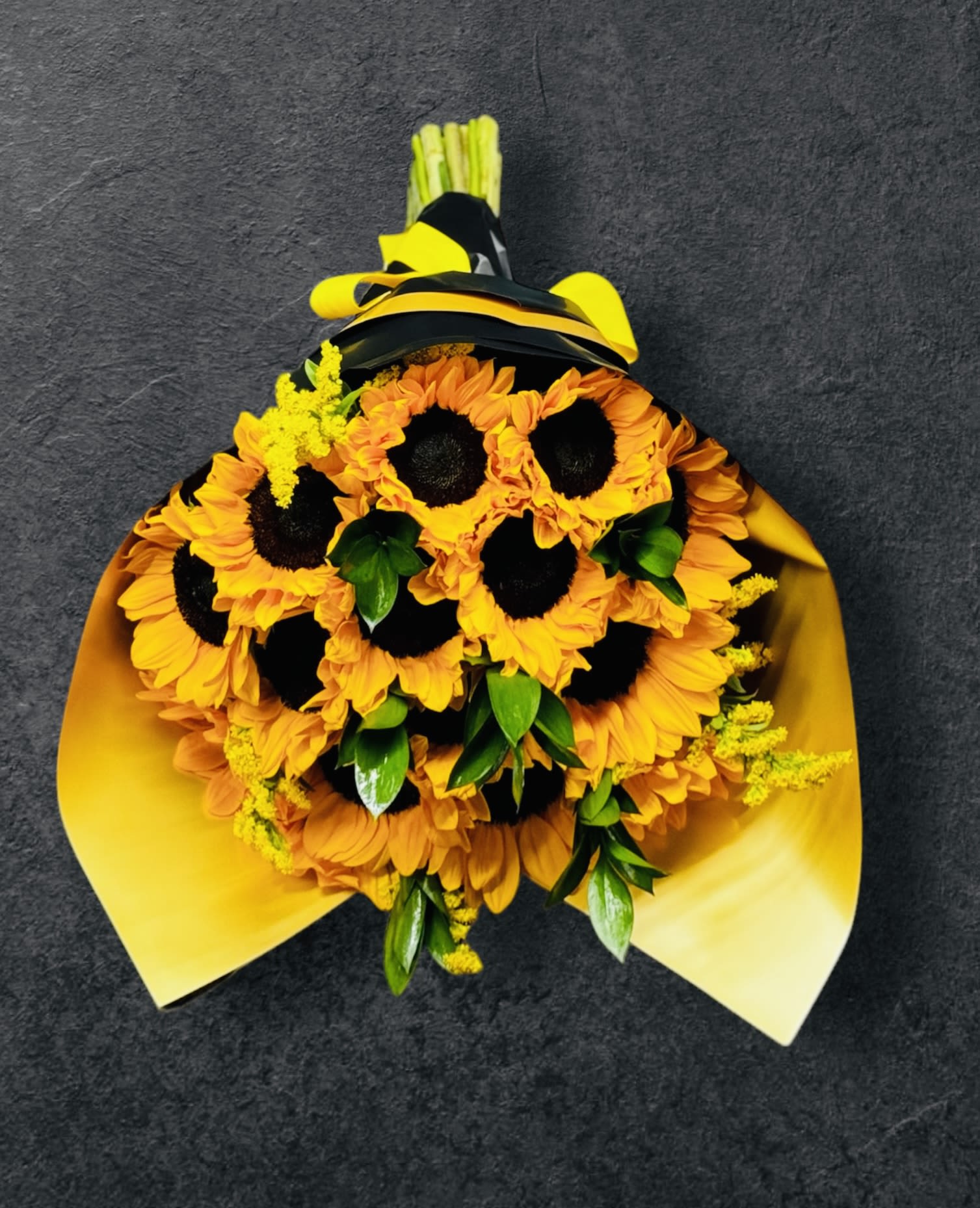 Bouquet of bright yellow sunflowers with green accents wrapped in yellow paper and black ribbon