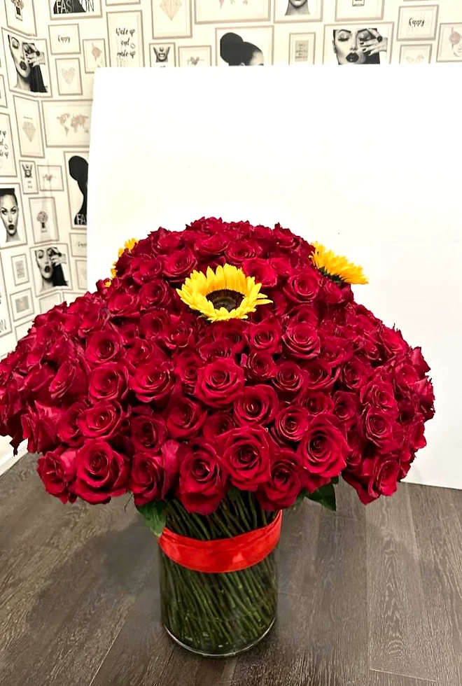 Large arrangement of red roses with yellow sunflowers in a clear glass cylinder vase with red ribbon.