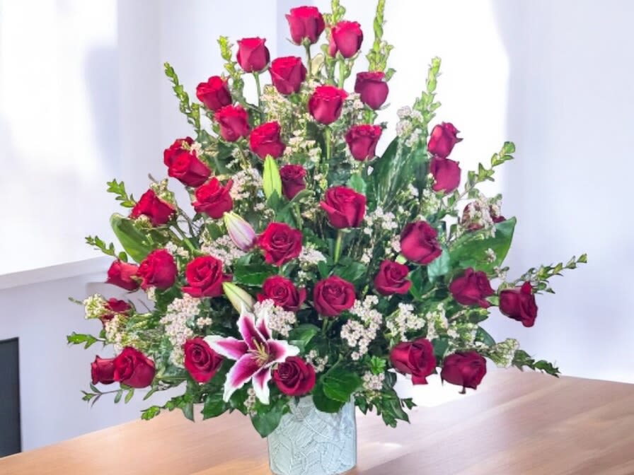 Tall arrangement of red roses with pink lilies and white filler flowers in a white ceramic vase