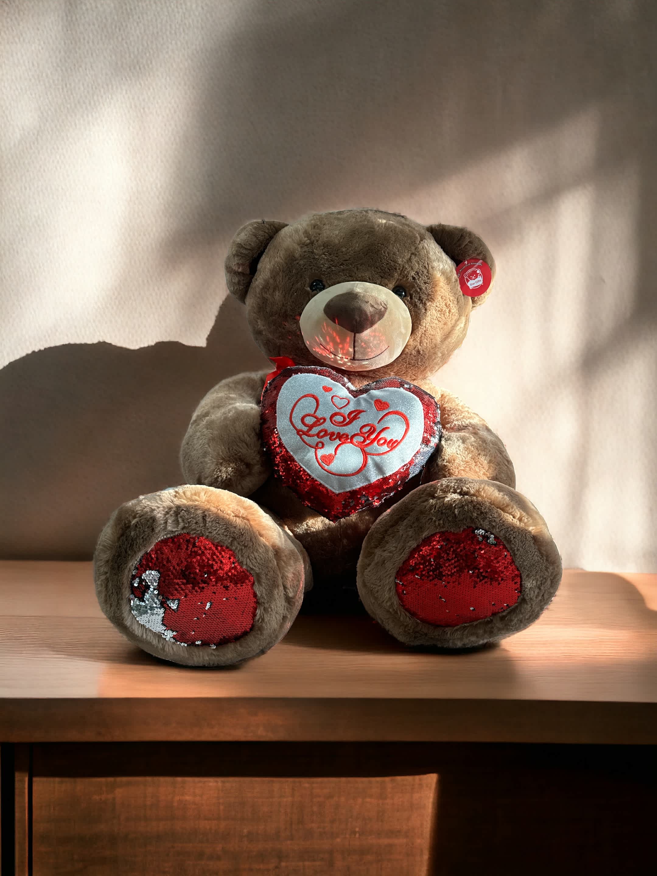 Brown teddy bear with red sequin heart reading "I Love You" on a wooden surface