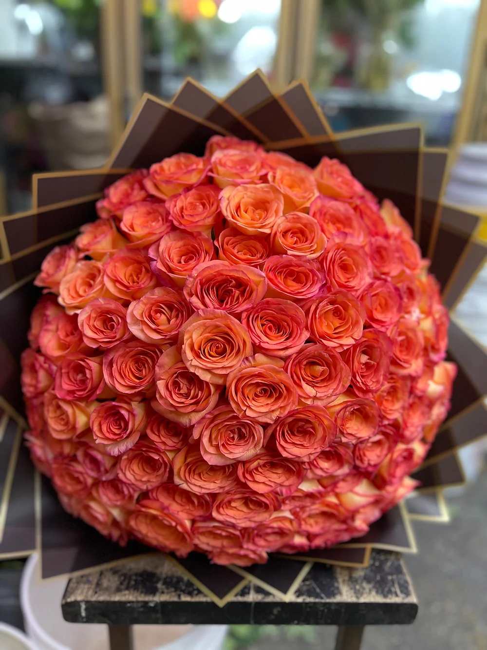 Large spherical bouquet of orange and pink roses wrapped in dark fan-folded paper.