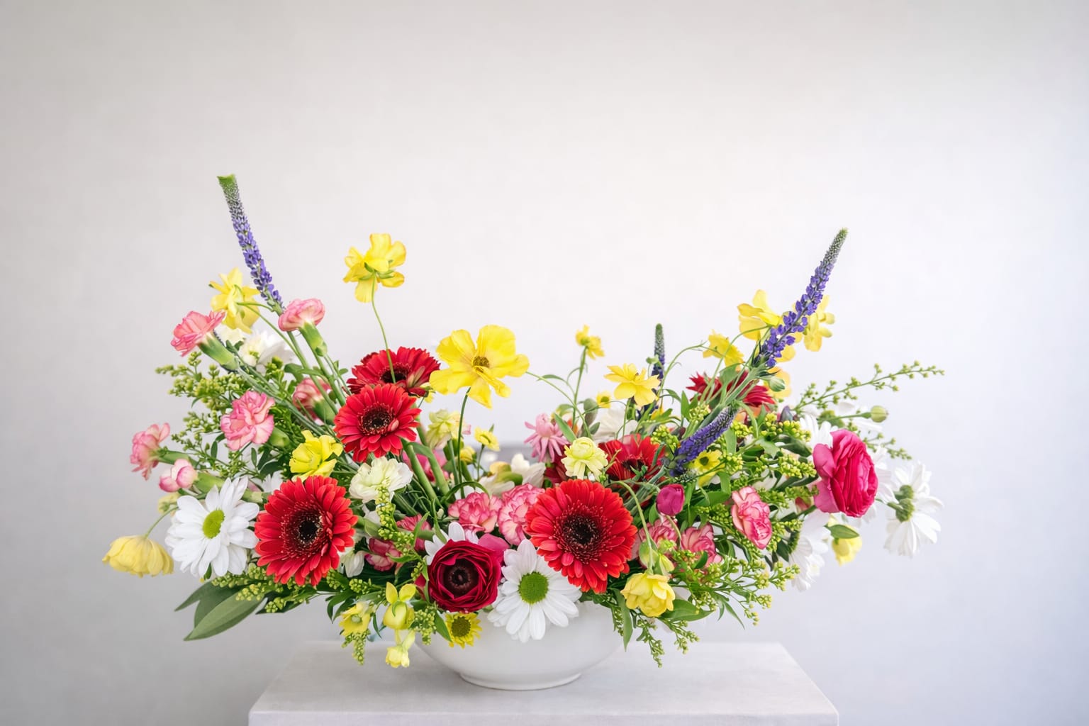 Sunlit Garden Symphony  - Vibrant red gerbera daisies, buttery yellow blooms, soft pink accents, and crisp white florals dance together in a lively, garden-inspired composition.  Delicate textures and airy stems create a sense of natural spontaneity, while the low, elongated design keeps the arrangement elevated and intentional. Pops of color are balanced with fresh greenery and subtle vertical elements, giving the piece dimension and rhythm from every angle.  Perfect for celebrations, dining tables, or brightening any space, this arrangement is both playful and polished—a true statement of floral artistry.