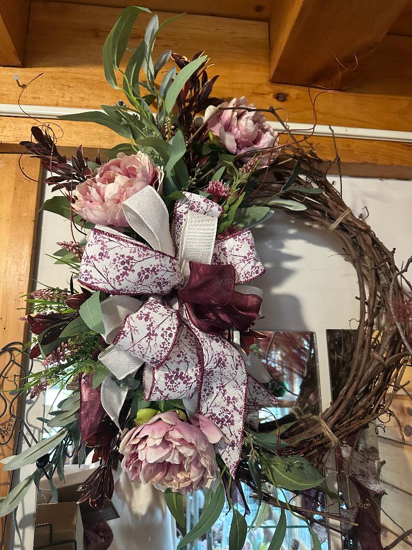 Grapevine wreath with pink peony blooms, greenery, and large white and burgundy patterned bow.