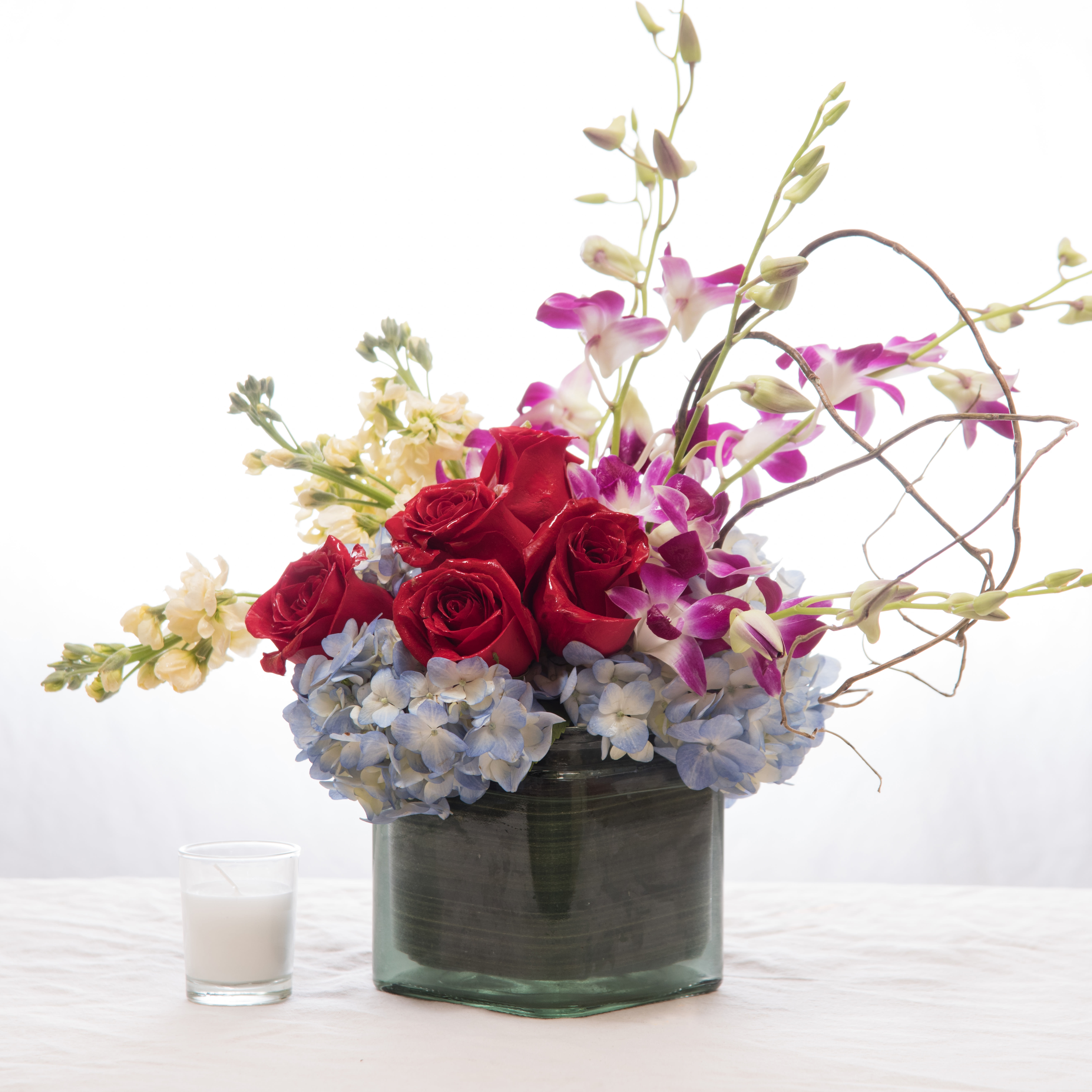Low arrangement of red roses, purple orchids, and blue hydrangeas in a glass vase with a small white candle.